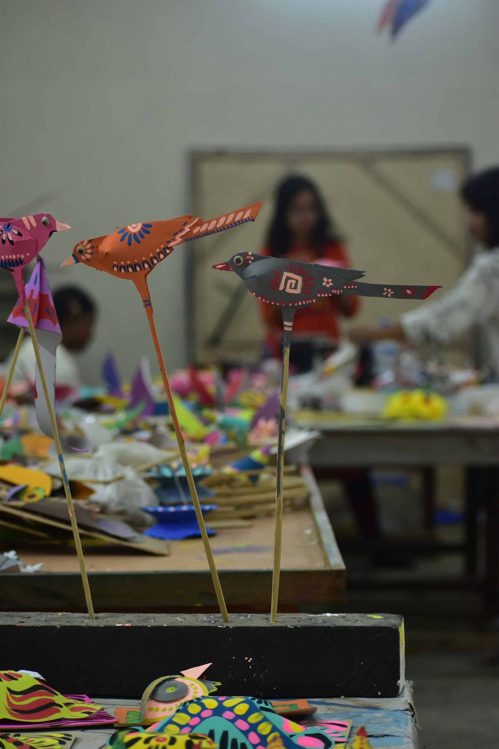 Colorful paper birds on sticks displayed on a table with arts and crafts supplies, with people in the background at an indoor craft event.