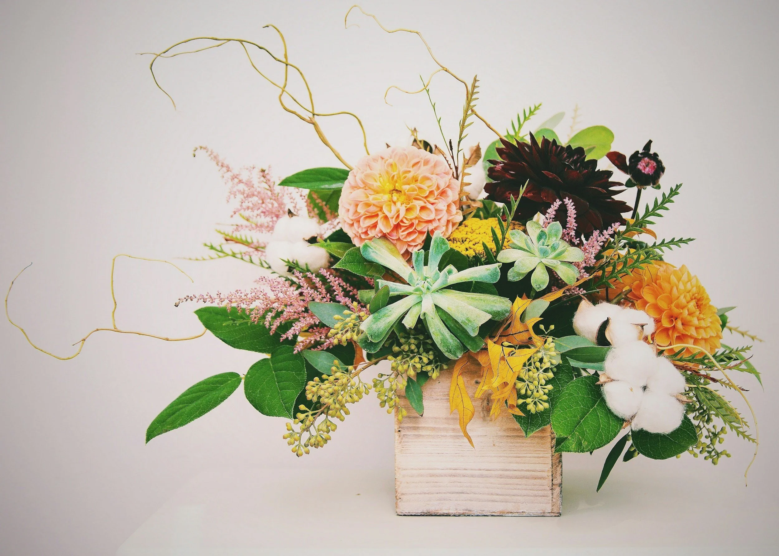 A mixed flower arrangement in a wooden box, including a pink dahlia, dark red dahlias, white cotton, pink astilbe, succulents, cotton stems, and green leaves, set against a plain light background.