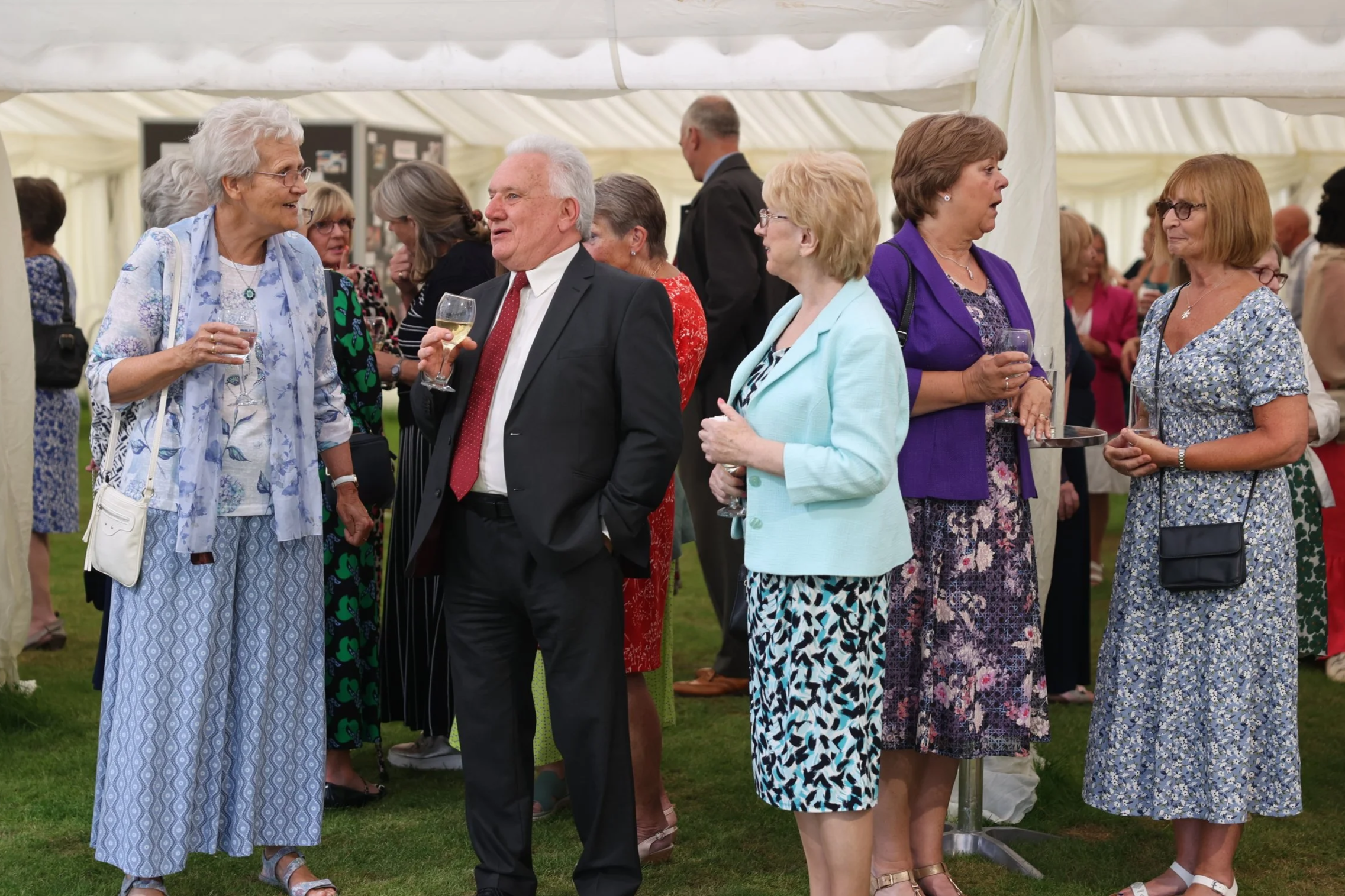 People dressed in colorful formal attire conversing at an outdoor event under a large white tent.