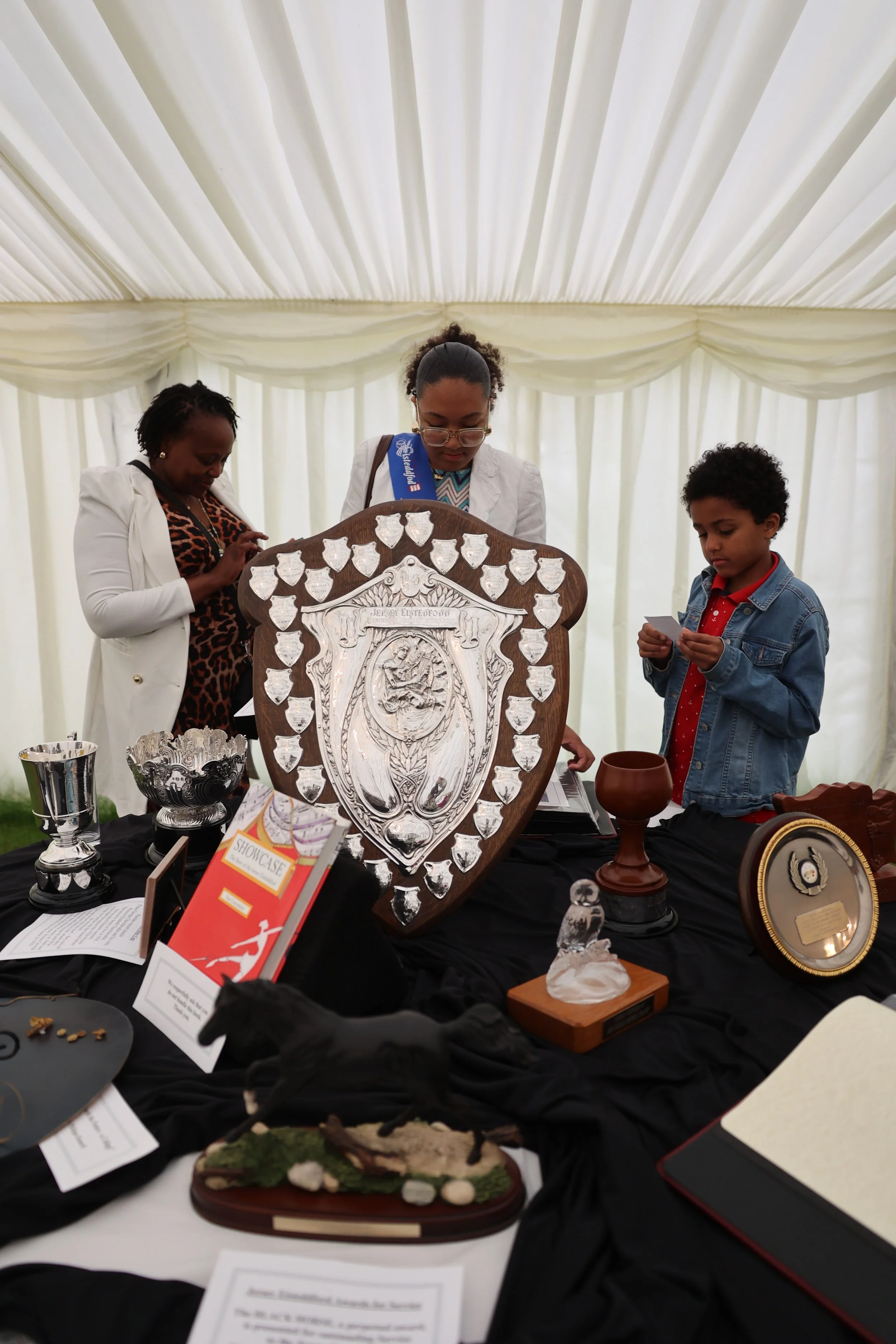 People examining awards and trophies at a display table under a white tent with draped fabric ceiling.