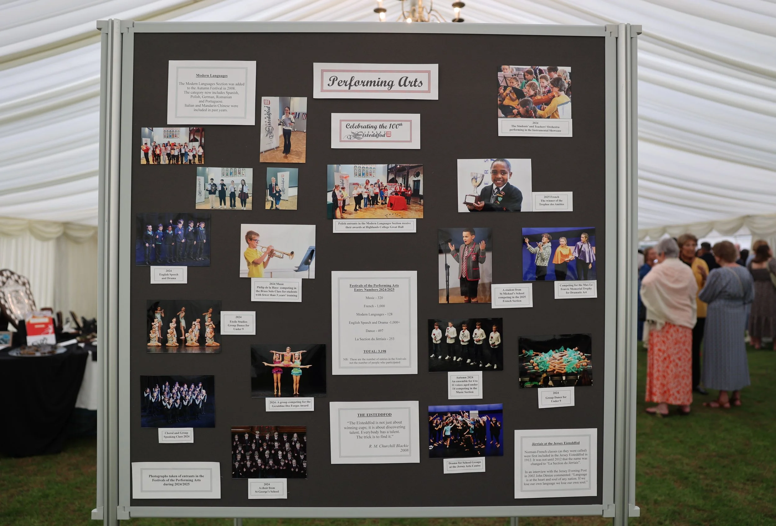A display board at a performing arts event showing photos and text about various performances and awards, with people visible in the background inside a tent.