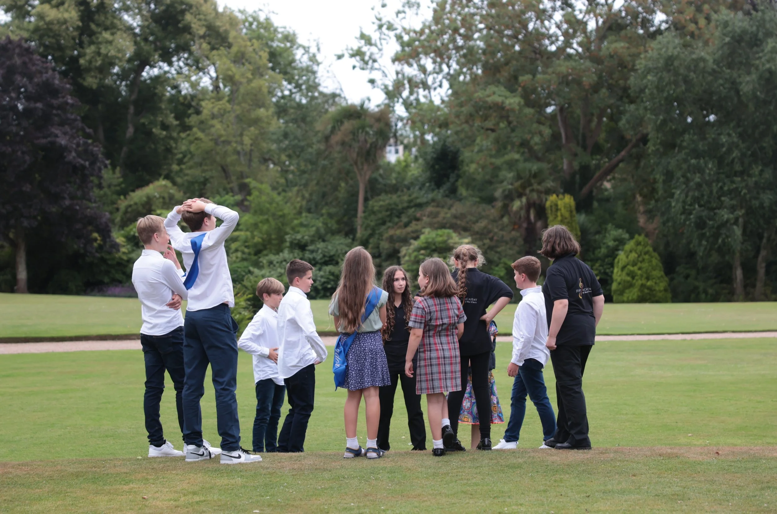 A group of children and teenagers gathered outdoors on a grassy field with trees in the background, engaging in conversation or activities.