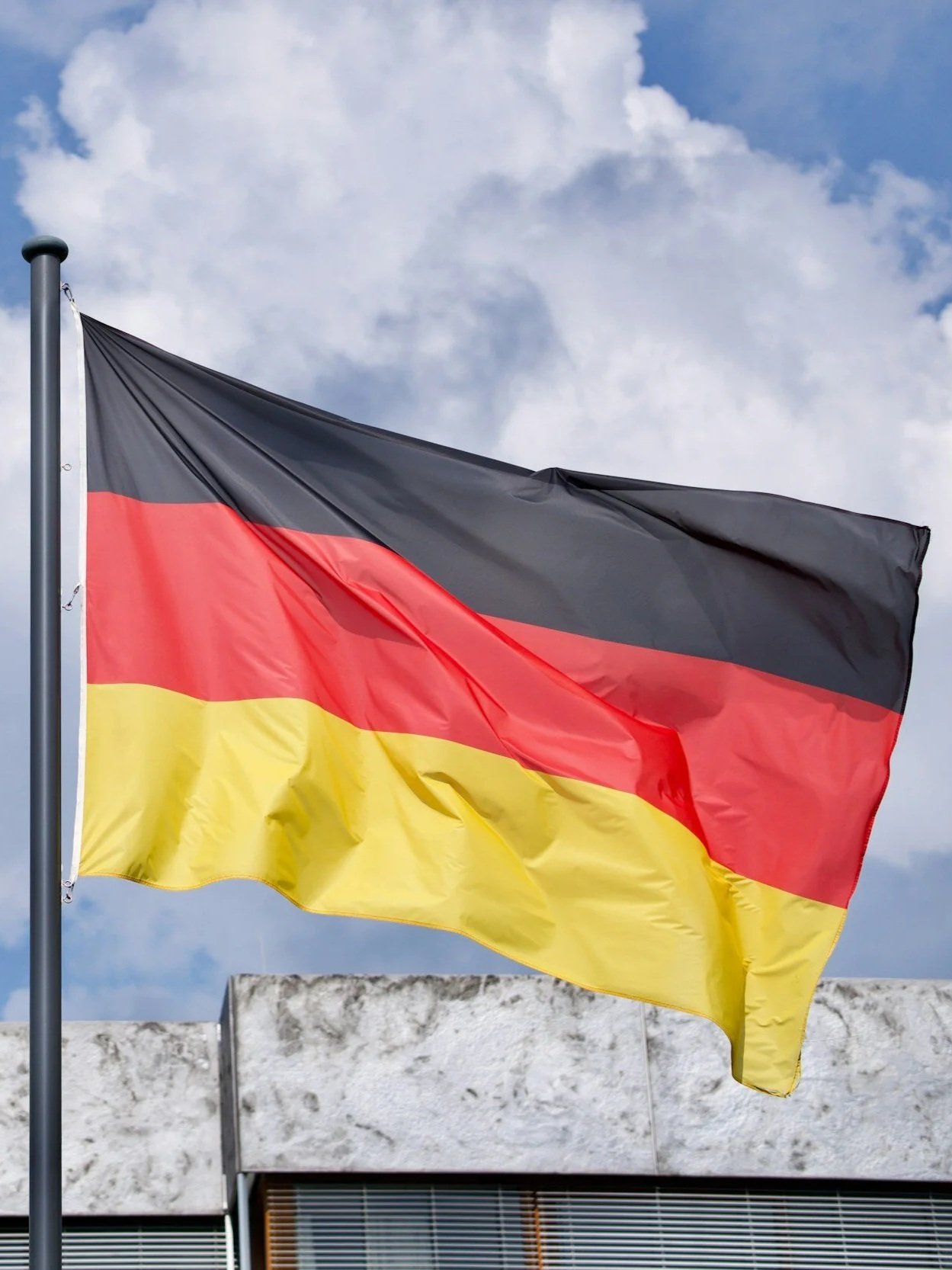German flag with black, red, and yellow horizontal stripes flying outdoors against a partly cloudy sky.