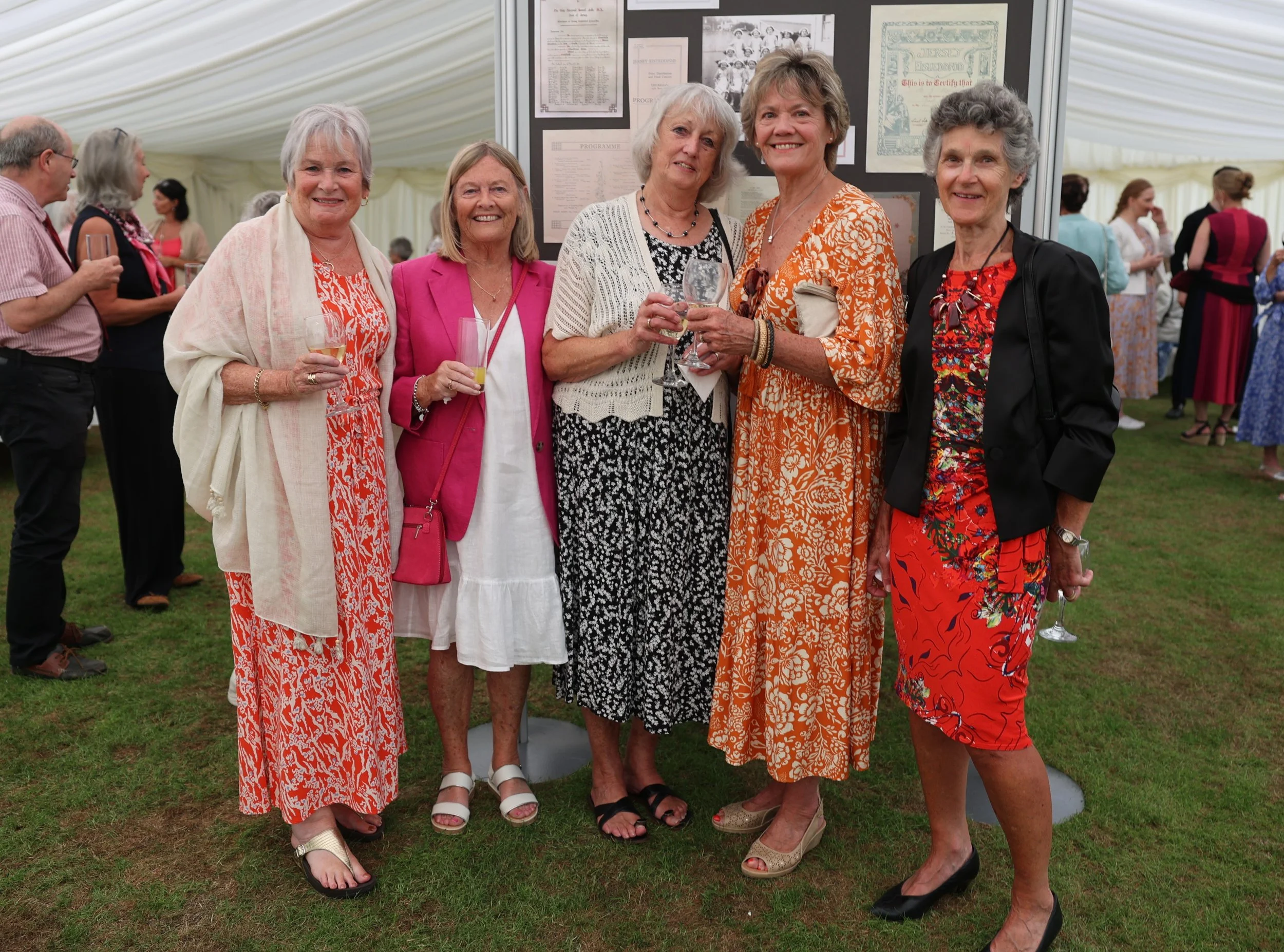 Group of six elderly women standing together at an event, holding glasses and smiling, with a display board behind them and other people socializing in the background, inside a large tent.