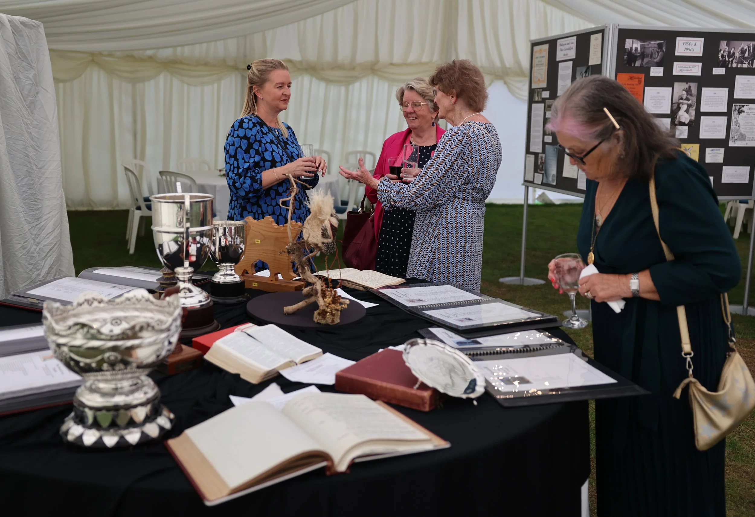 Women at an outdoor exhibition or fair browsing display tables with books, artifacts, and informational posters under a tent.