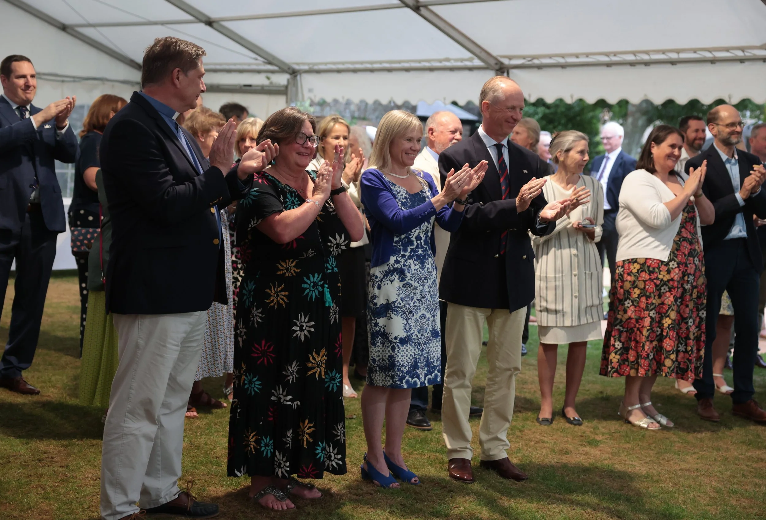 A group of people in formal and semi-formal attire standing close together inside a tent, clapping and smiling at an event.