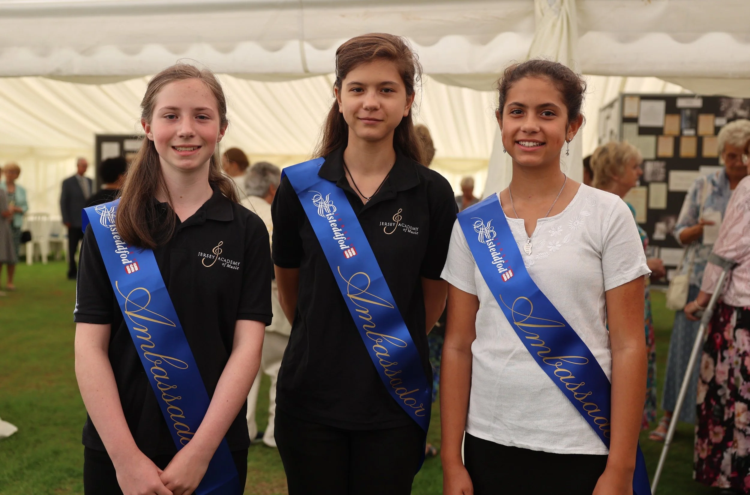 Three young girls wearing sash that say 'Congratulations' standing inside a large tent during a celebration.