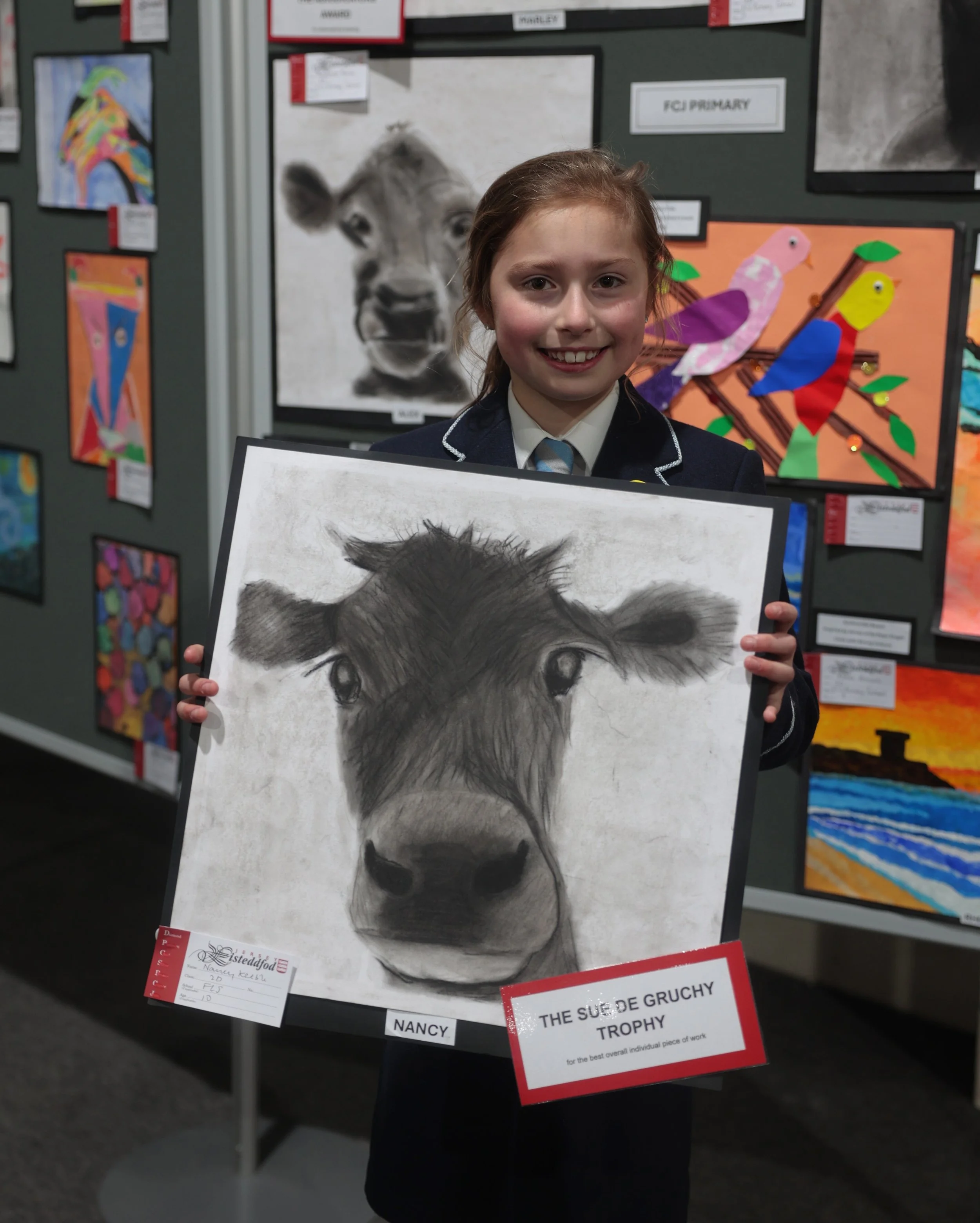 A young girl dressed in a formal school uniform holding a black and white pencil drawing of a animal's face, likely a wolf or a dog, at an art exhibition. She has long, reddish hair and is smiling.