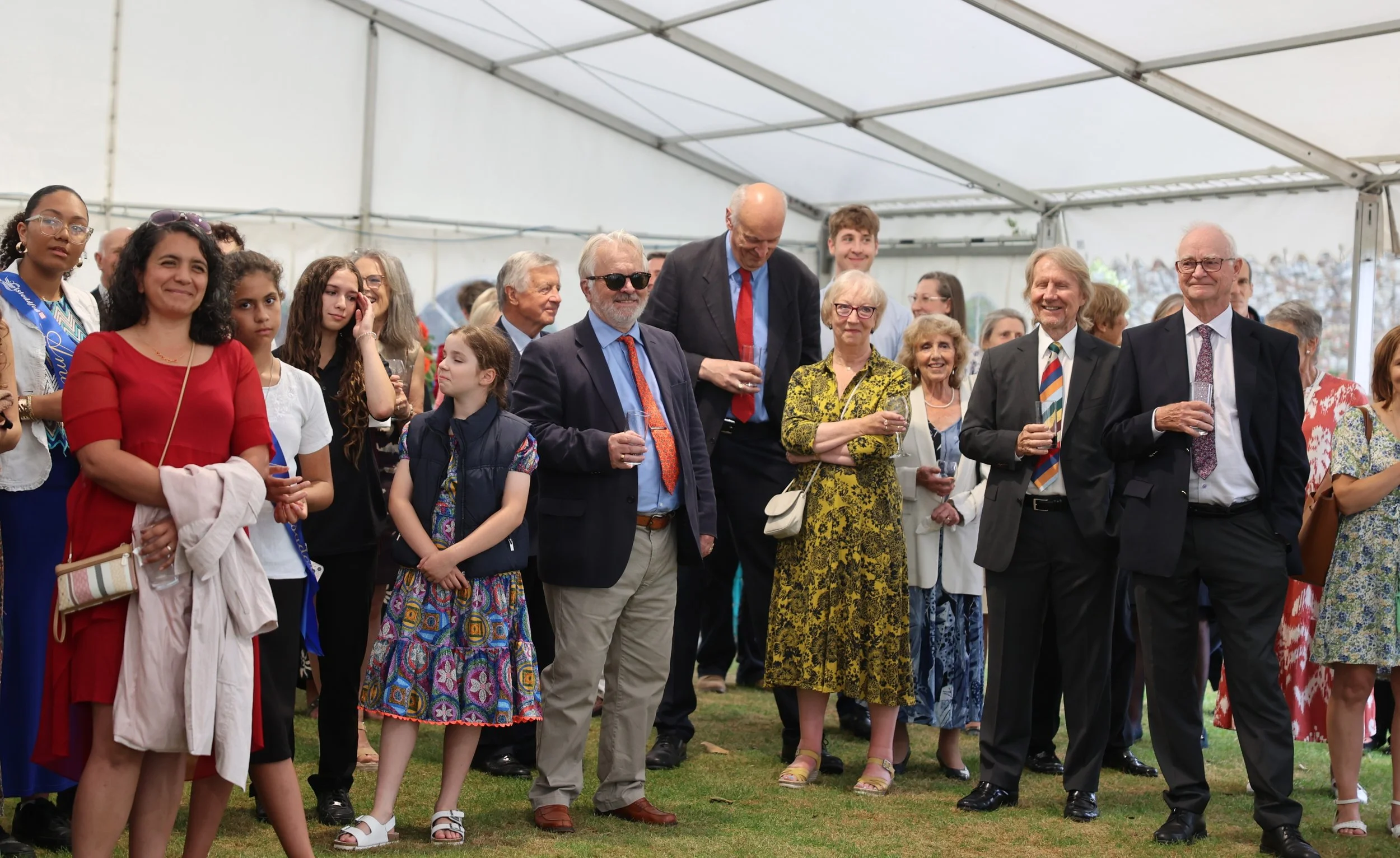 A diverse group of people, including children and elderly, gathered under a large tent at a celebration or event, with some holding glasses of drinks and smiling.