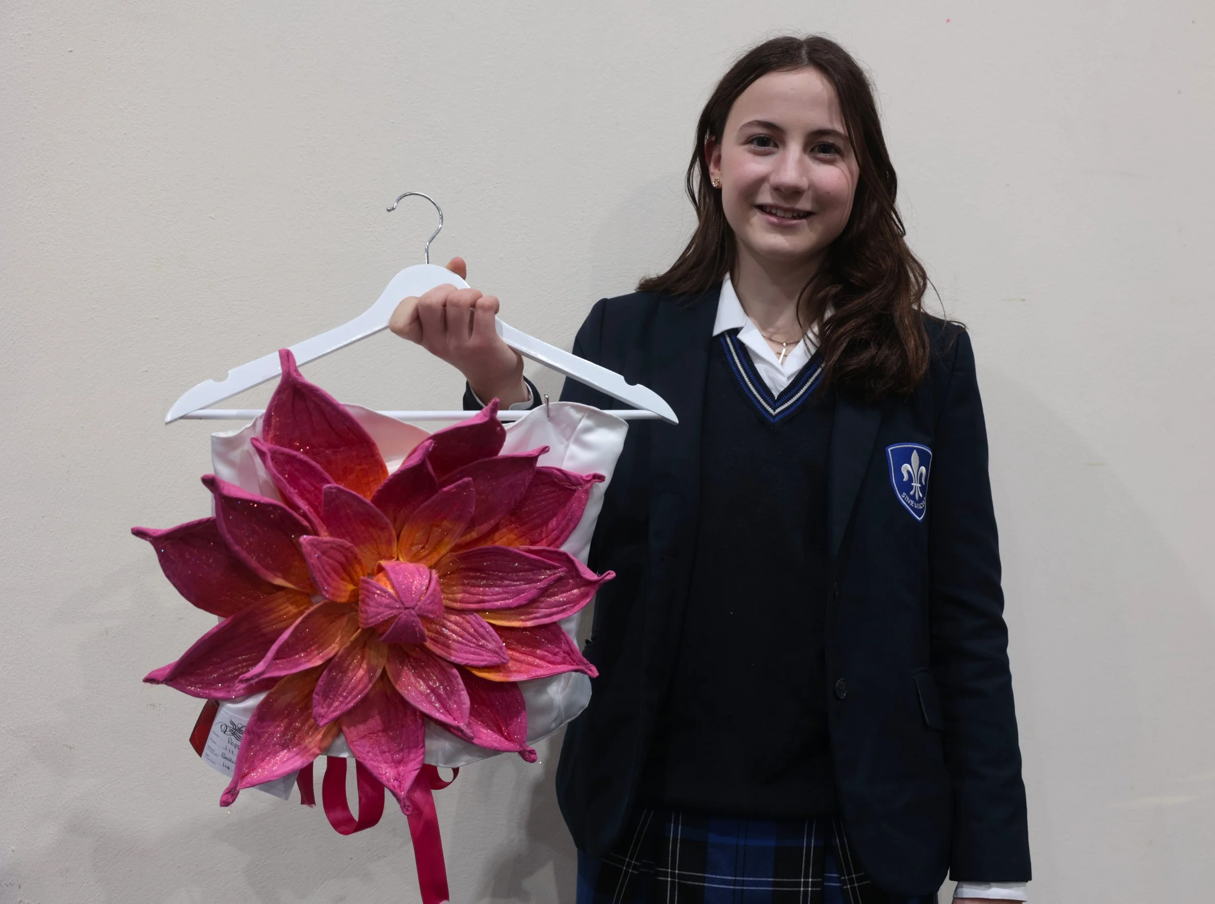 A young girl wearing a navy blazer with a school badge, smiling, holding a white hanger with a large pink flower-shaped decoration attached to it, standing against a plain studio background.