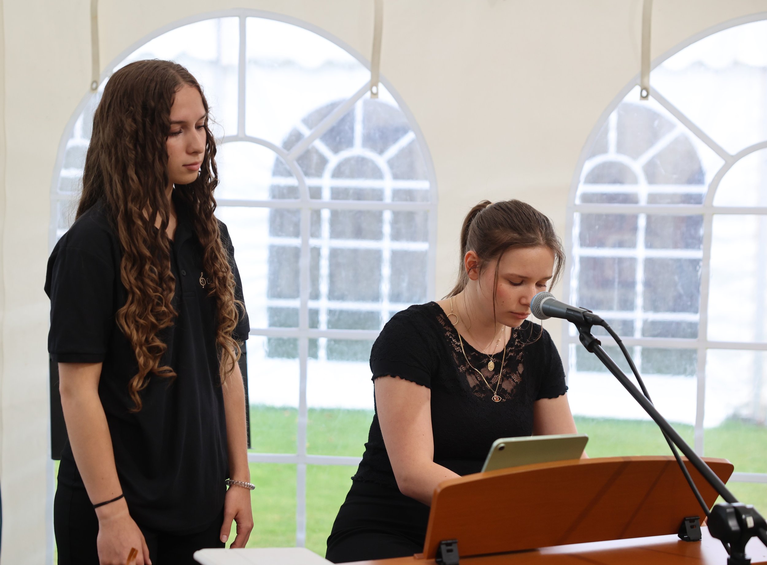 Two young women are performing music indoors, one is singing while the other is playing the piano.