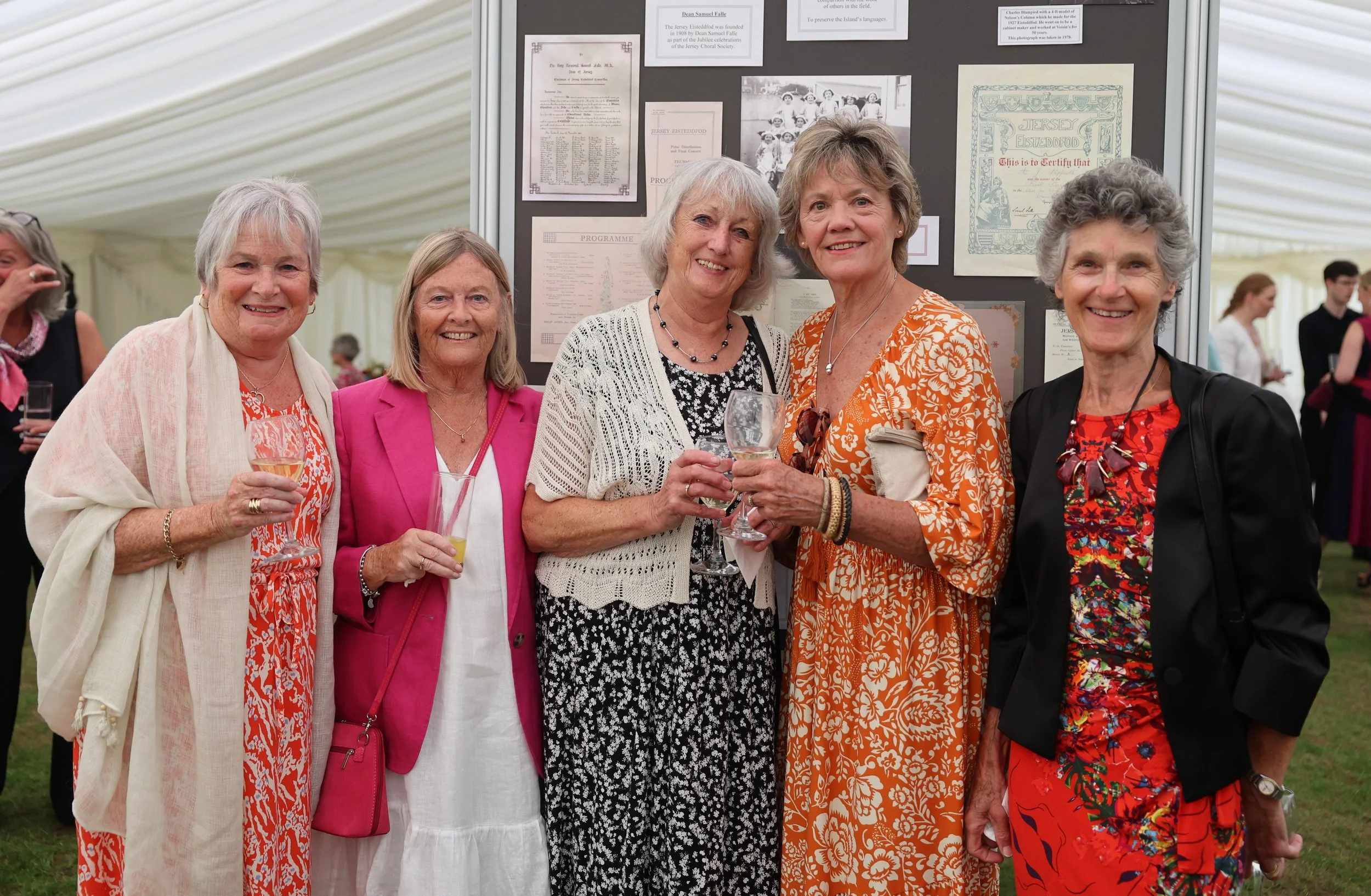 A group of six older women standing together at a social event inside a tent, smiling, some holding glasses of wine, with a display board with photos and documents behind them.
