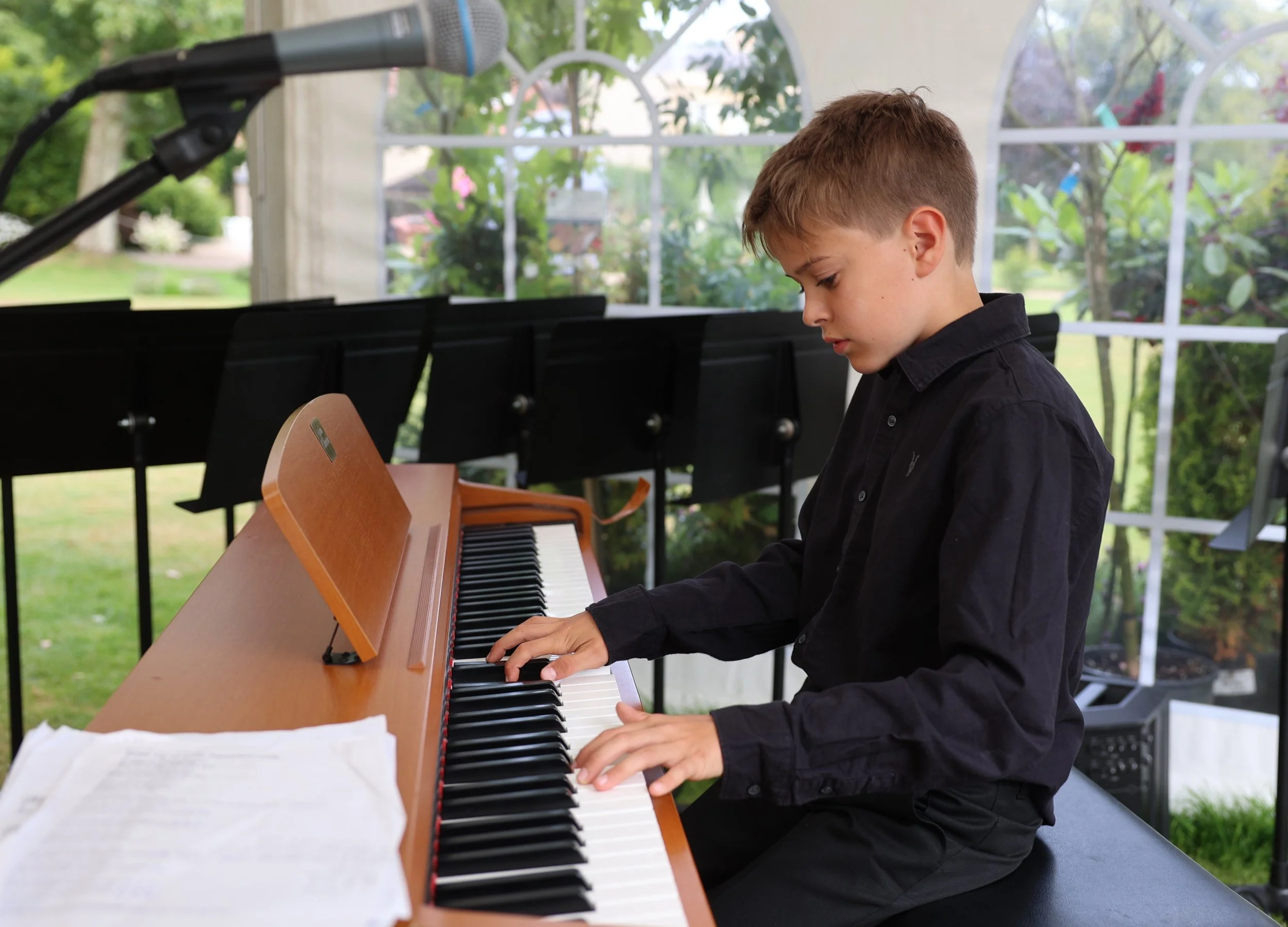 A young boy playing a wooden upright piano outdoors, with sheets of music and a microphone nearby, in a bright greenhouse or conservatory surrounded by greenery.