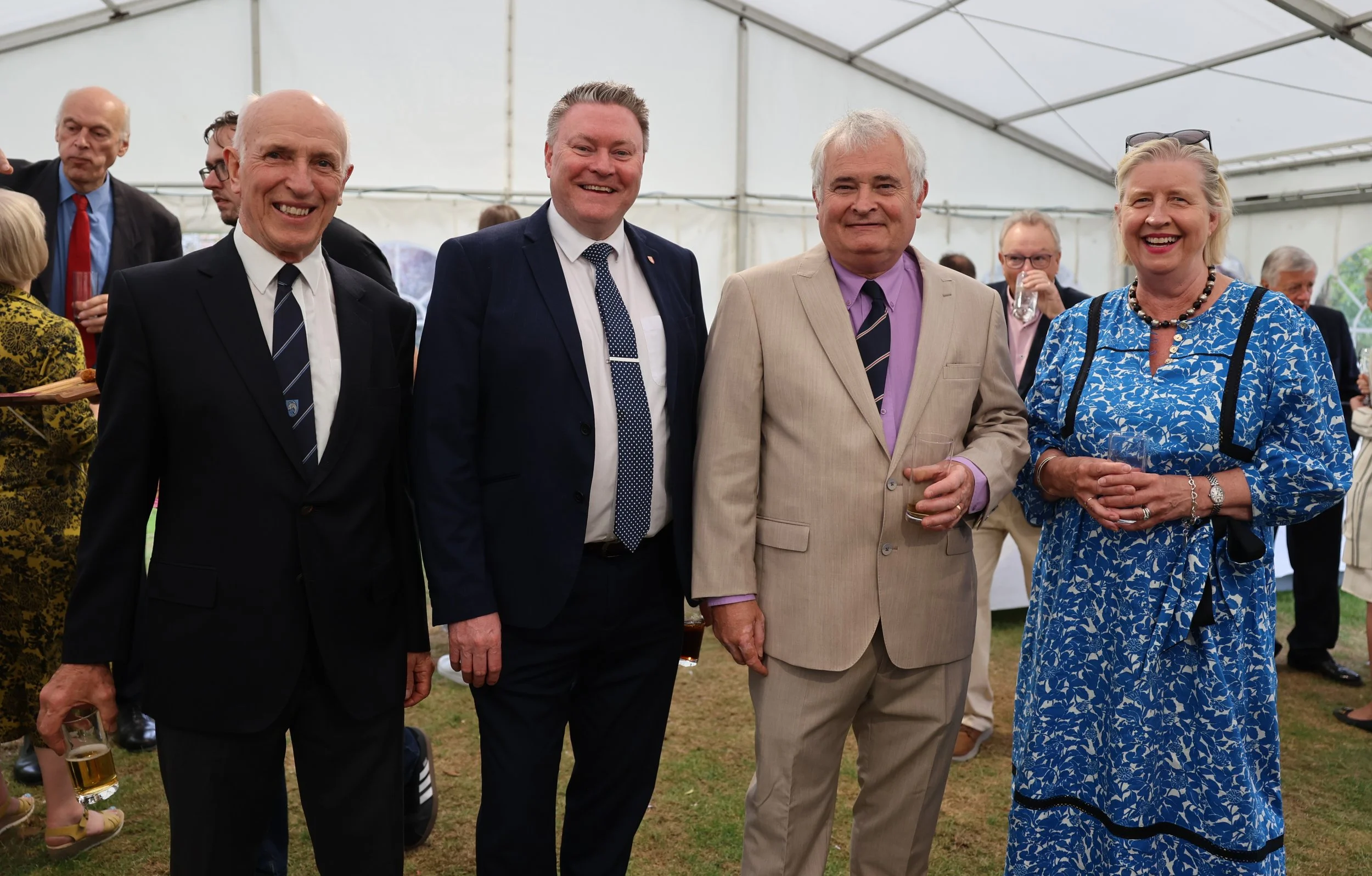 Group of four people standing together in a tent during a social event, holding drinks, smiling, dressed in formal and semi-formal attire.
