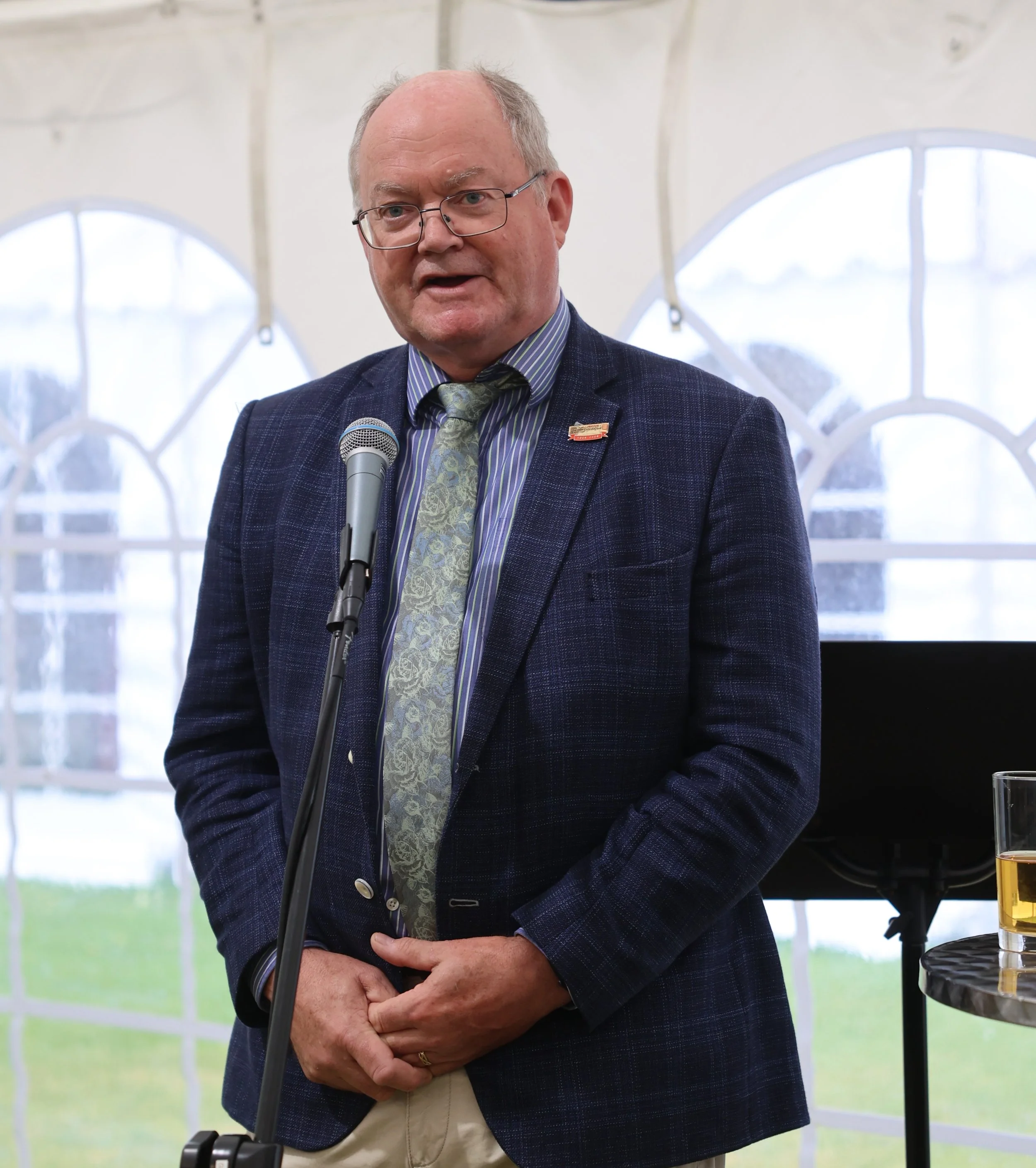 A bald man wearing glasses, a dark blue blazer with a pin, a striped shirt, and a tie, speaking into a microphone at an indoor event with large windows showing a snowy landscape outside.