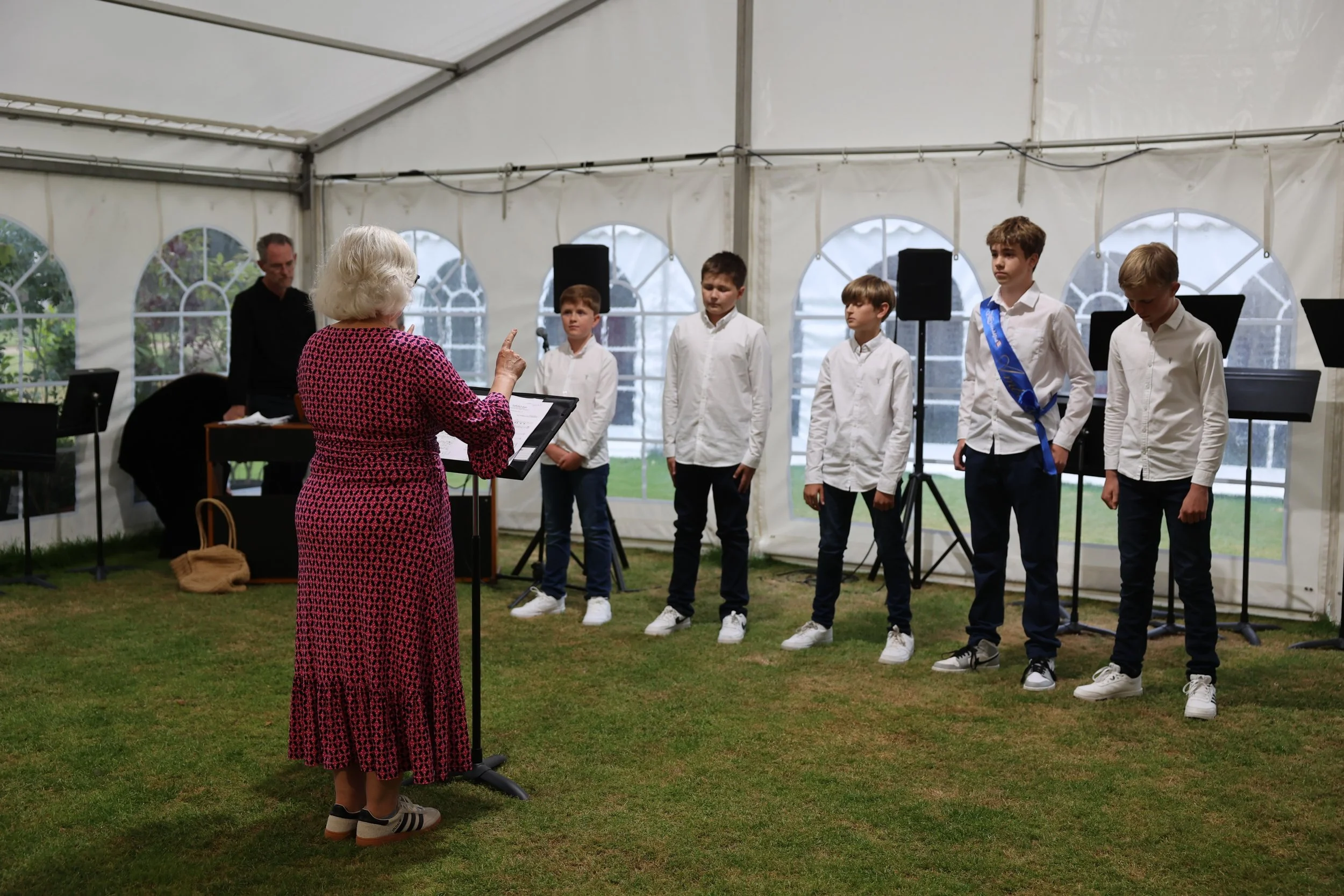 An elderly woman in a pink and black dress conducting a choir of five young boys, five of them have white shirts and black pants, inside a white event tent.