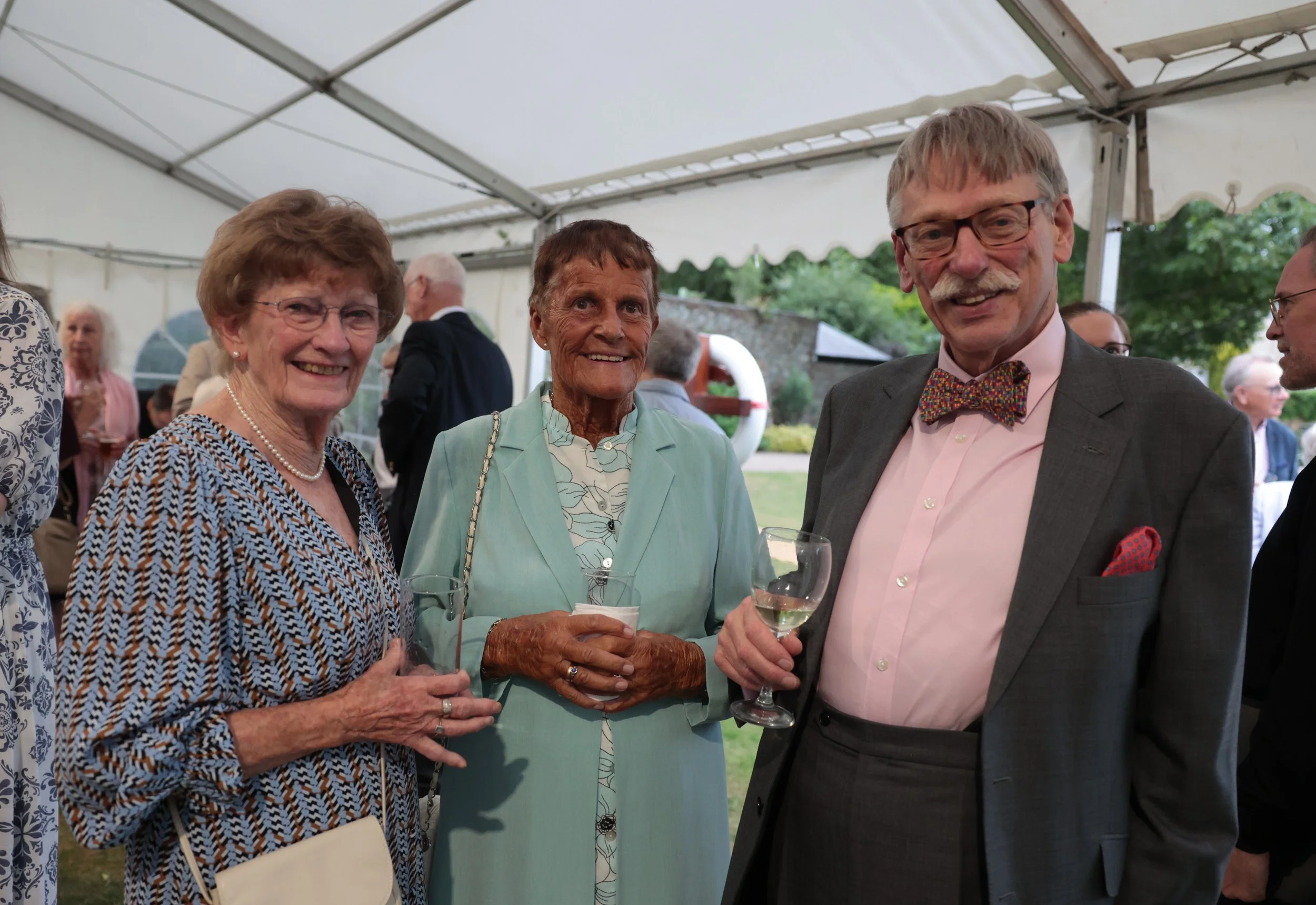 Three elderly people in formal attire smiling and holding wine glasses at a social event under a tent.
