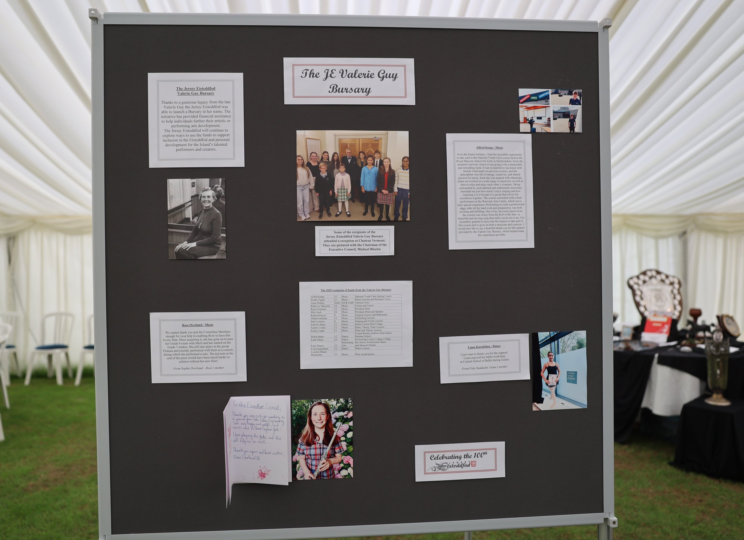 A large black display board shows various photos and articles about Valerie Guy Bursary and other artists, displayed inside a white tent with chairs visible in the background.
