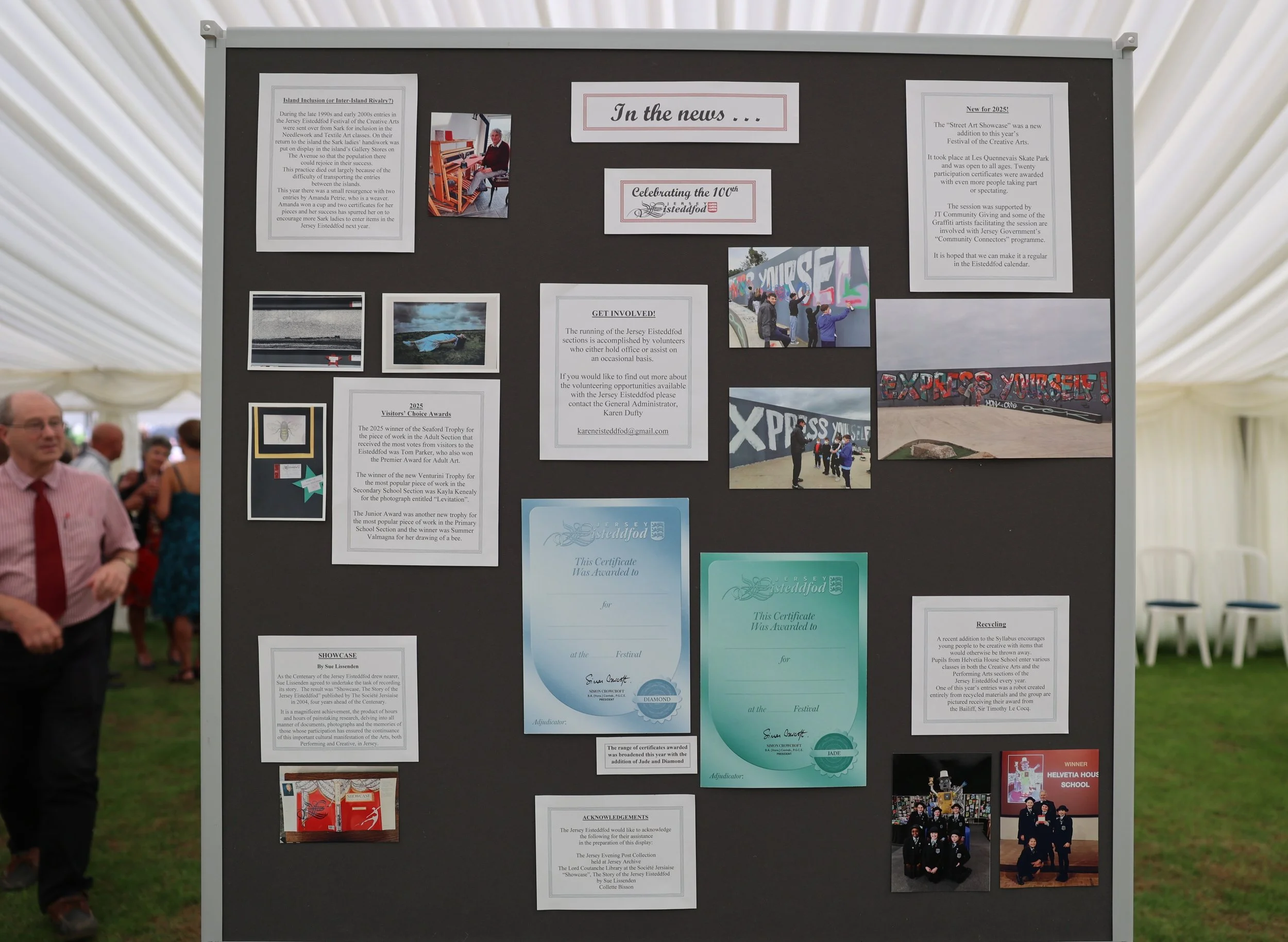 Black display board with various informational posters, images, and certificates about Jersey Eisteddfod, decorated with photos of people, graffiti art, and awards, set in an outdoor tent at a festival event.