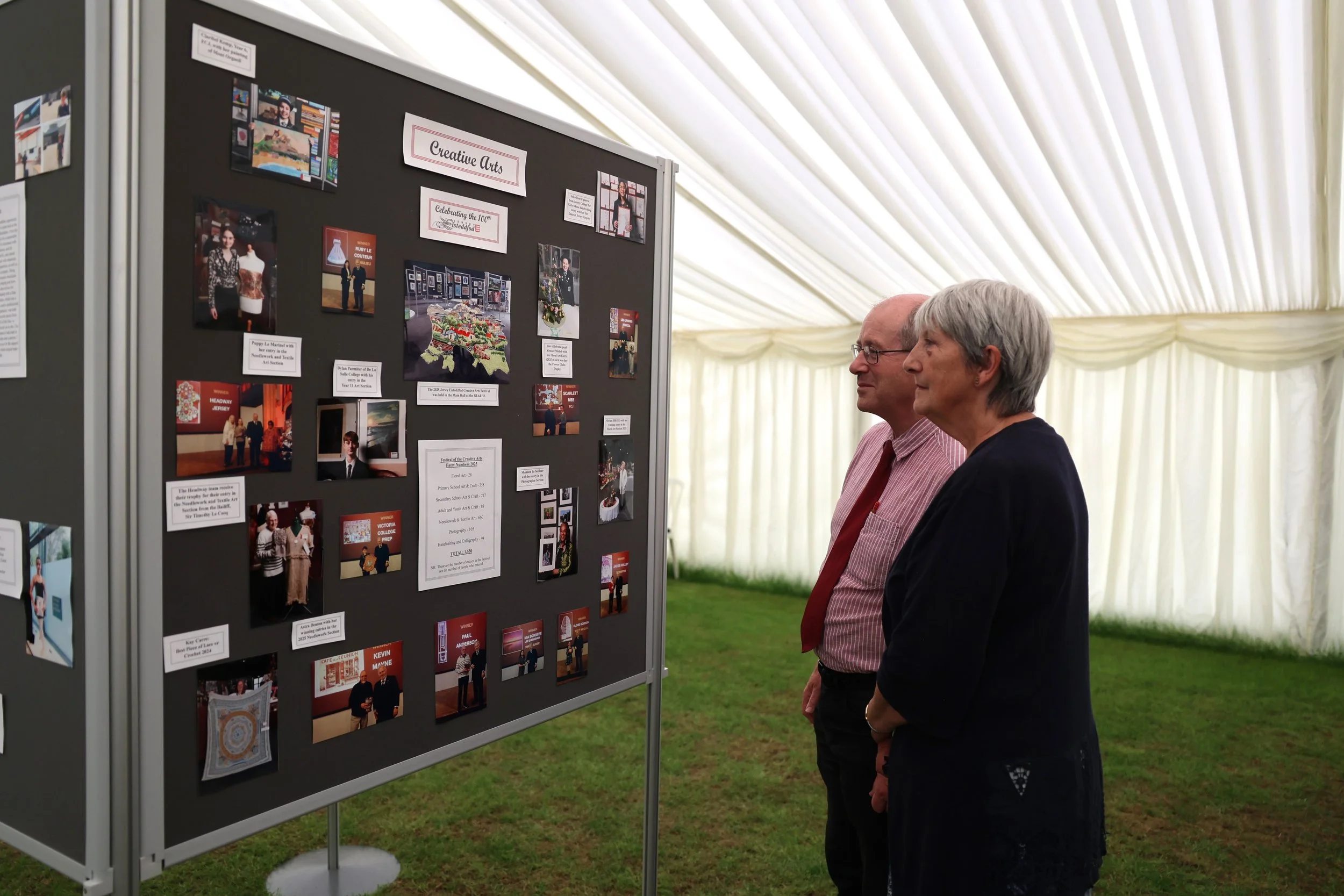 Two people, a man and a woman, are standing and observing a display board with photos and information at an event under a large white tent. The display board is titled 'Creative Arts'.