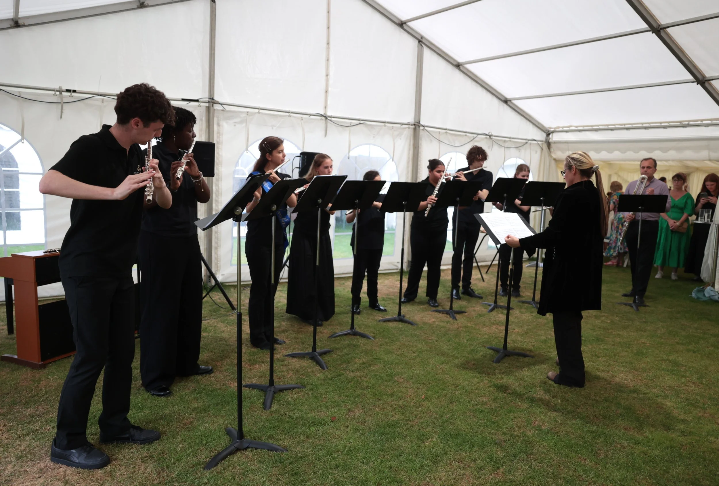 A group of young musicians performing under a large white event tent, with a conductor leading them and an audience in the background.