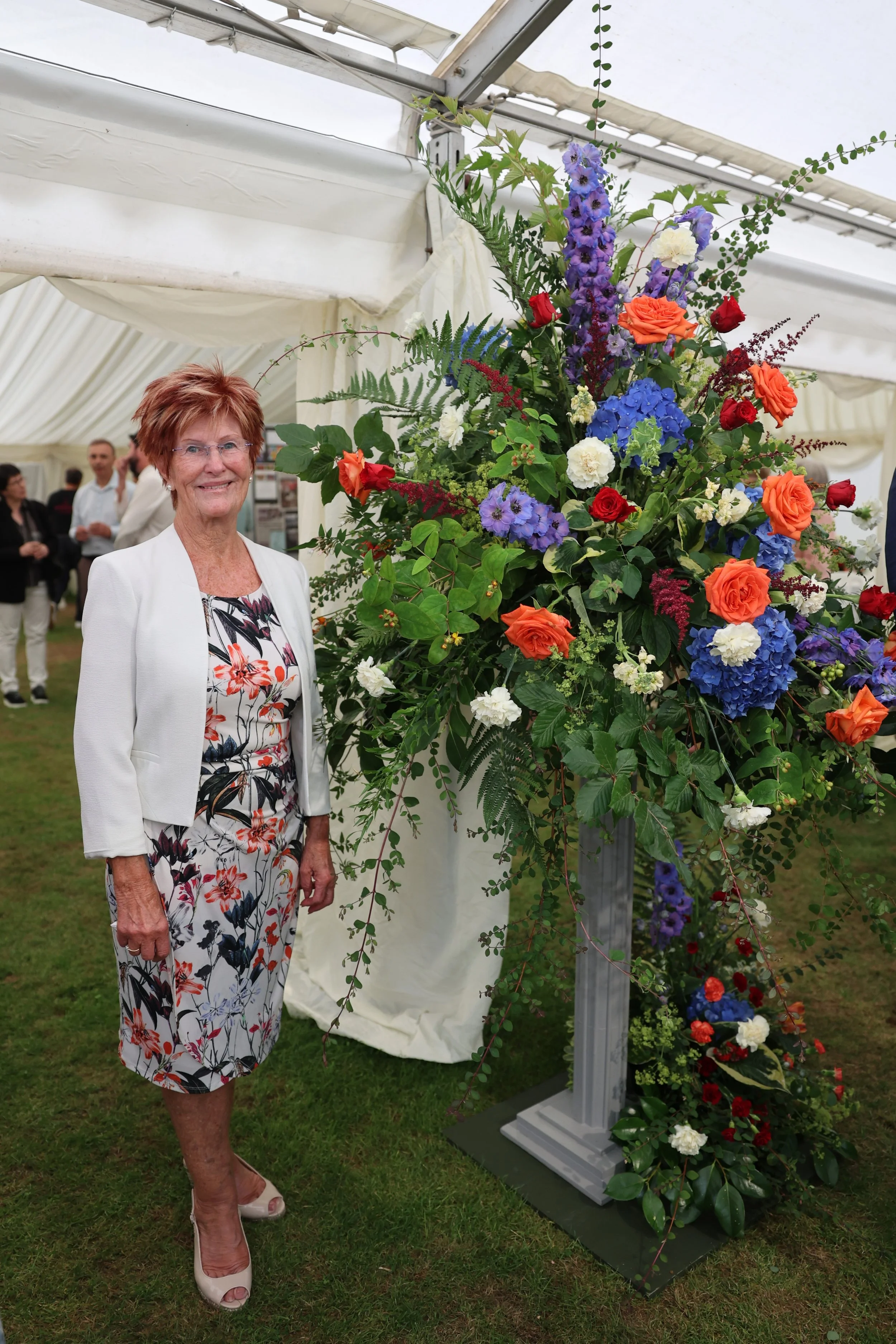 A woman in a floral dress and light-colored blazer stands next to a large colorful flower arrangement in a greenhouse or event tent, with other people in the background.