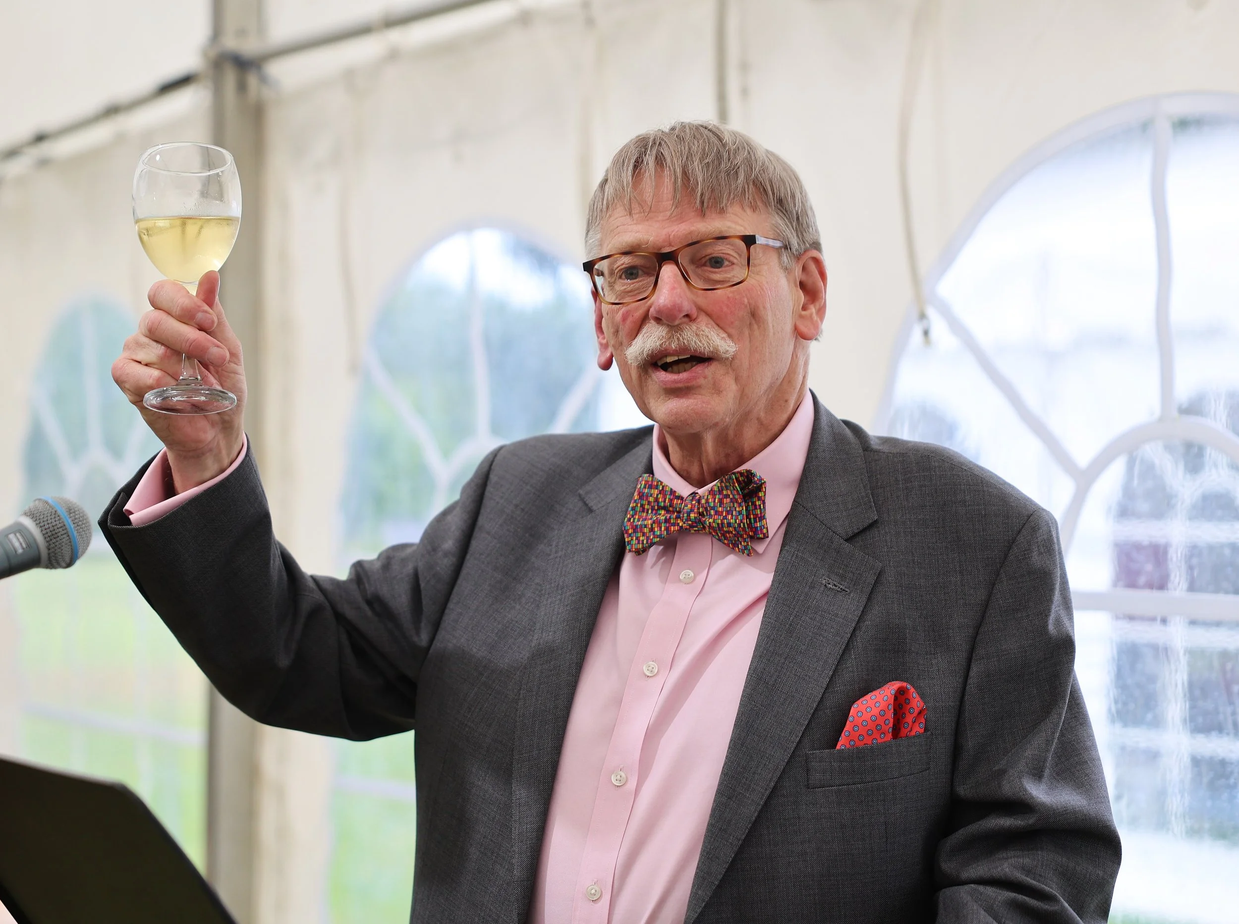 An elderly man with gray hair and glasses wearing a gray suit, pink shirt, and colorful bow tie, holding a glass of white wine in his right hand, speaking at an event.