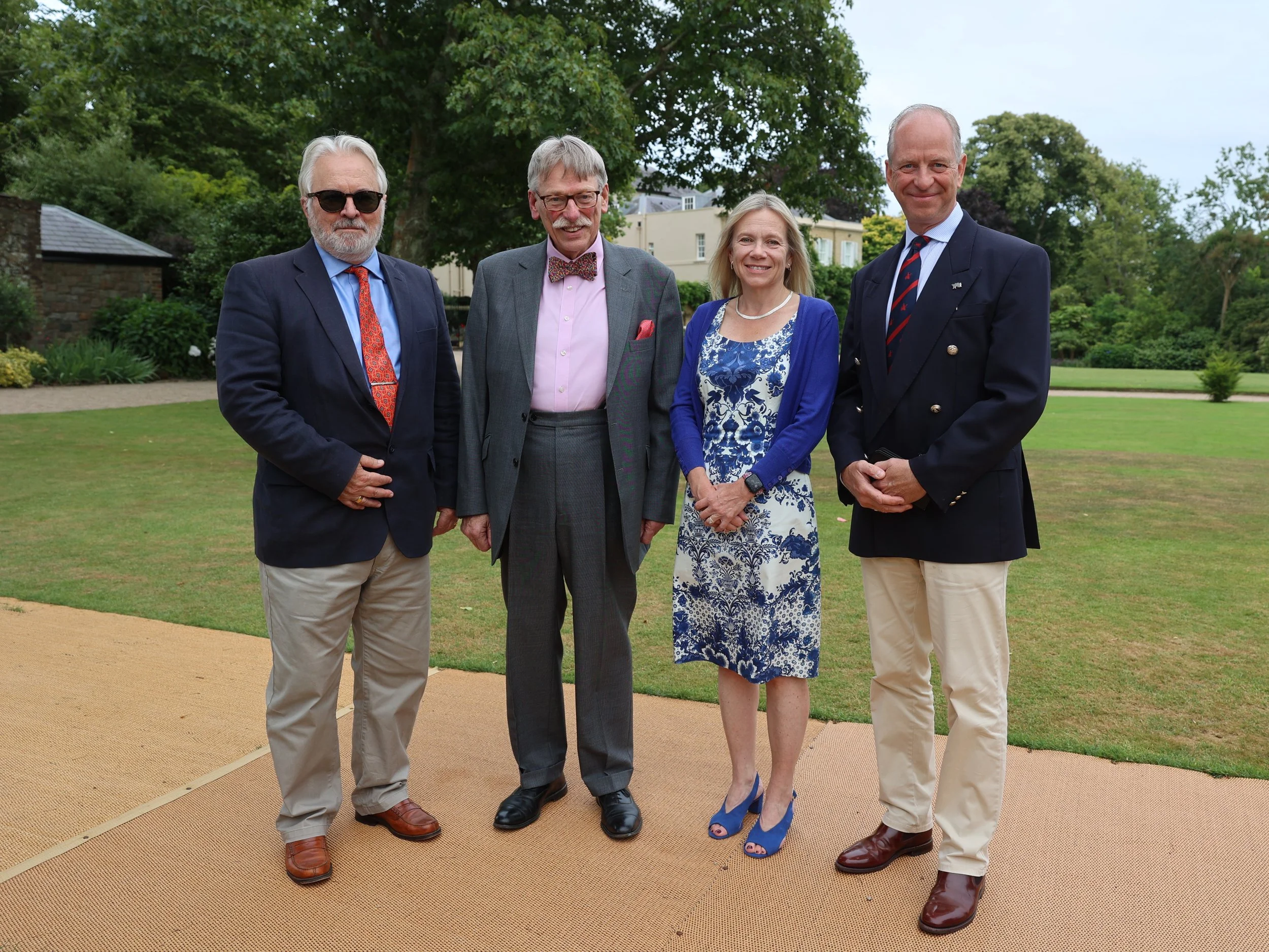Four elegantly dressed older adults standing outdoors on a lawn with trees and houses in the background, smiling at the camera.