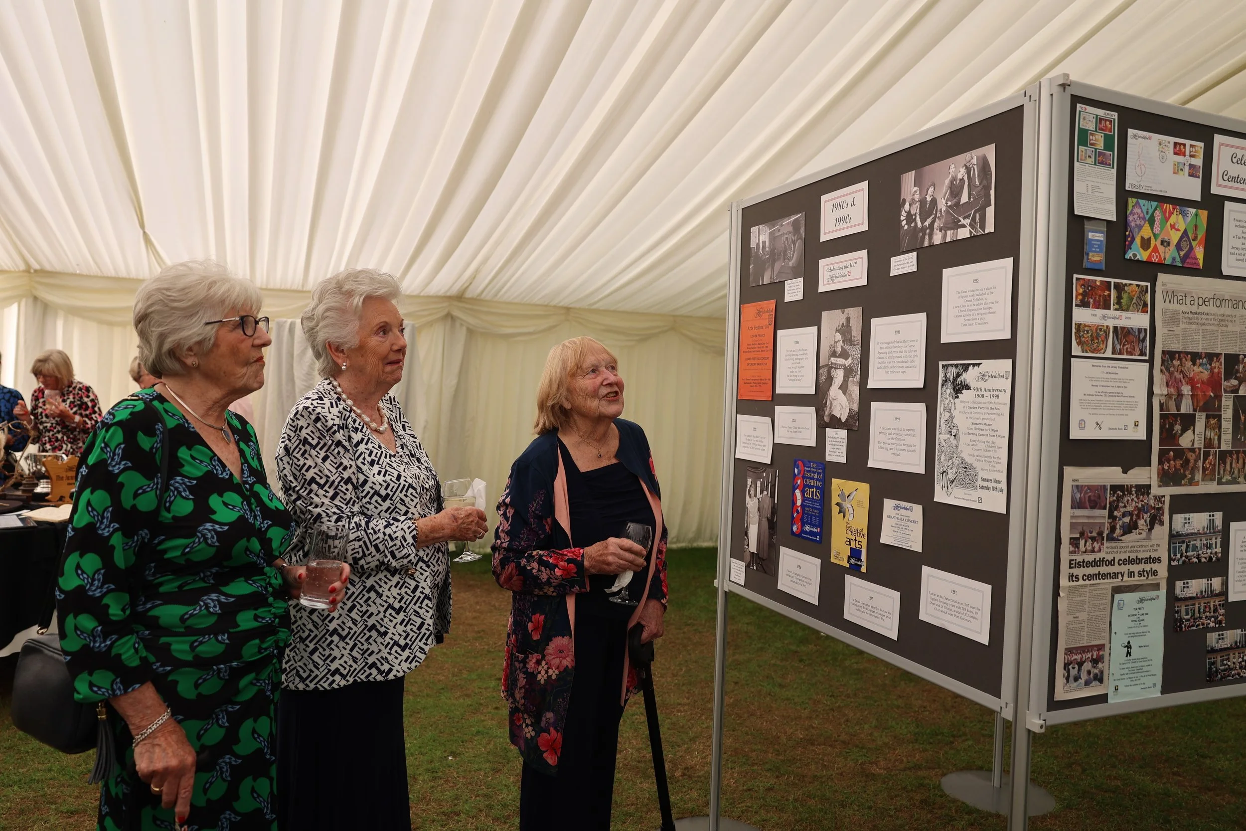Three elderly women standing inside a large event tent, looking at a display board with photos and information, holding drinks, one woman in a green patterned dress, another in black and white geometric, and the third in a dark floral dress.