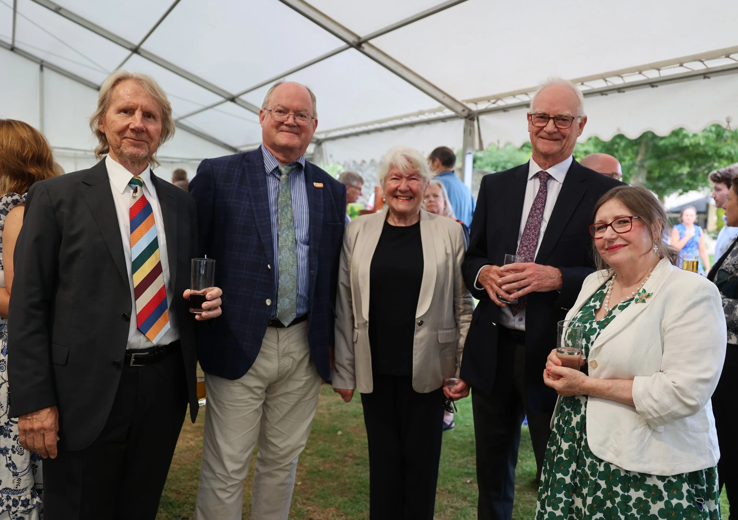 Group of six older adults at an outdoor event under a white tent, holding drinks and smiling at the camera.