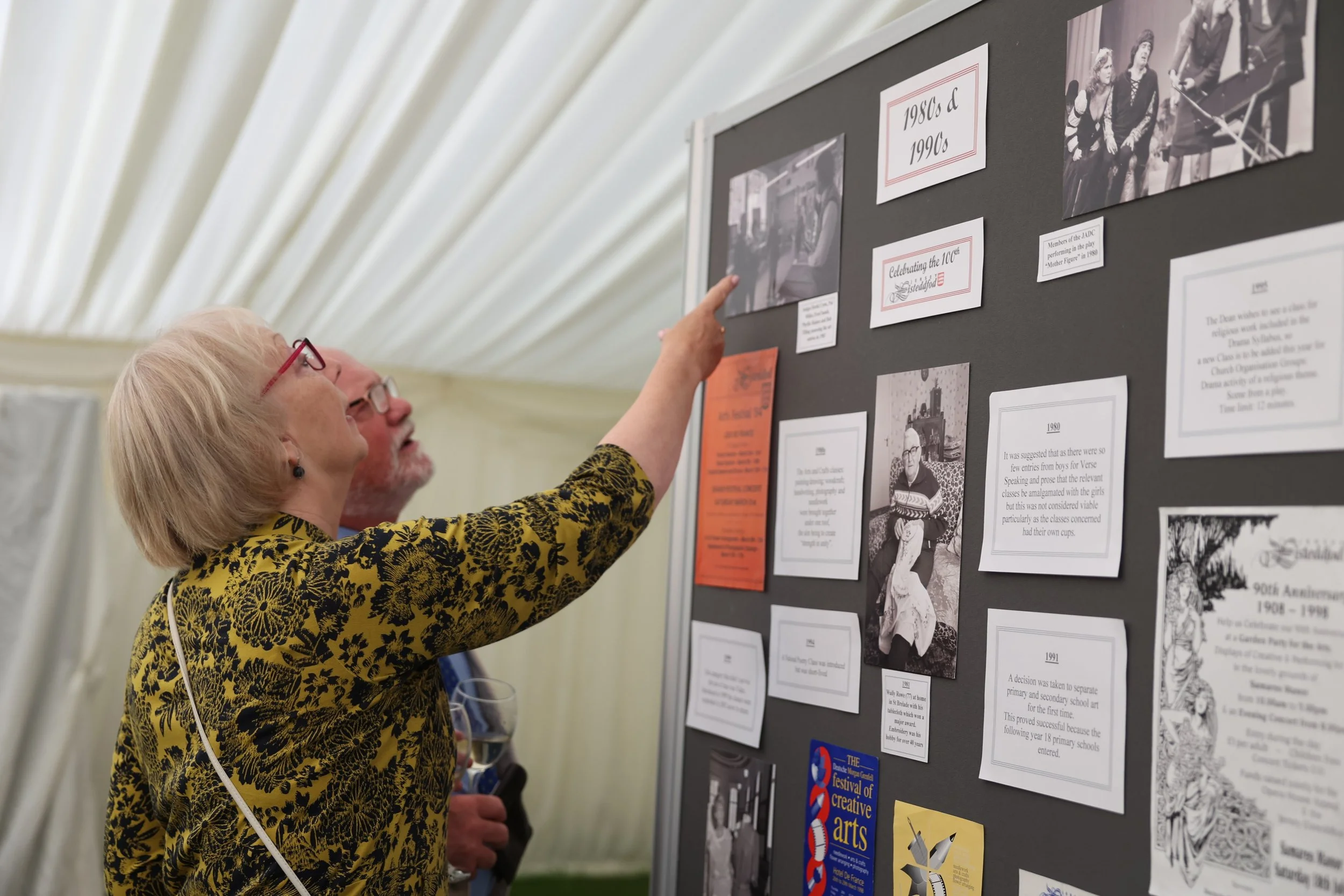 An elderly woman with blonde hair and glasses is pointing at a display board with black and white historical photographs and information, under a tent with a white, fabric ceiling.