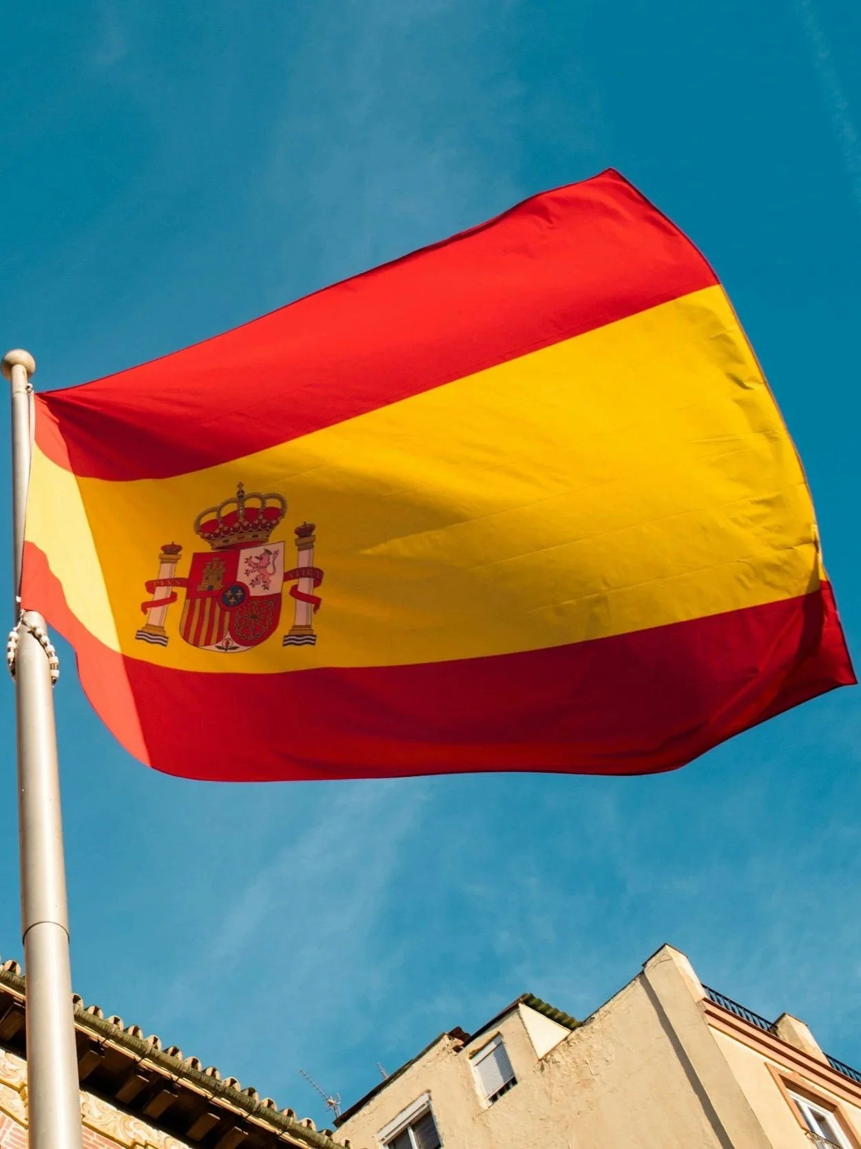 Spanish flag flying outdoors against a clear blue sky with buildings below.