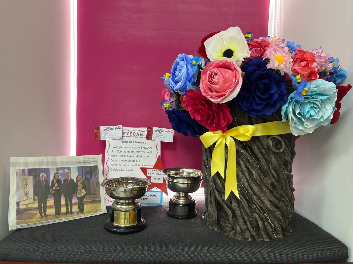 A memorial display featuring a large artificial bouquet of paper flowers in a wooden vase with a yellow ribbon, two trophies, a framed photo of four people in formal attire, and a sign that reads "Held in Memory" and mentions the 80th Liberation Celebration 2025.