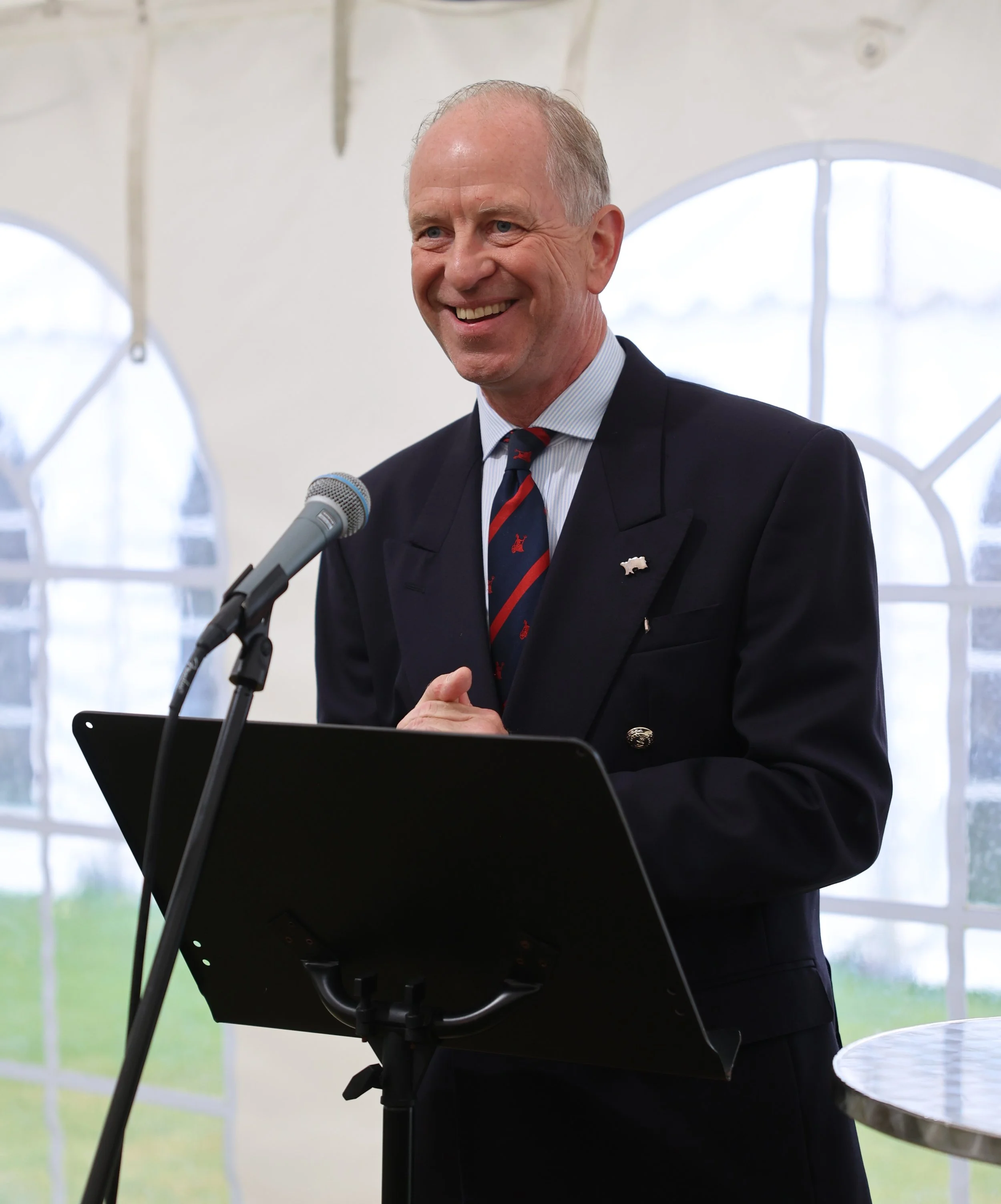 A man with a broad smile, dressed in a suit, standing in front of a white tent with round windows, speaking into a microphone during a formal event.