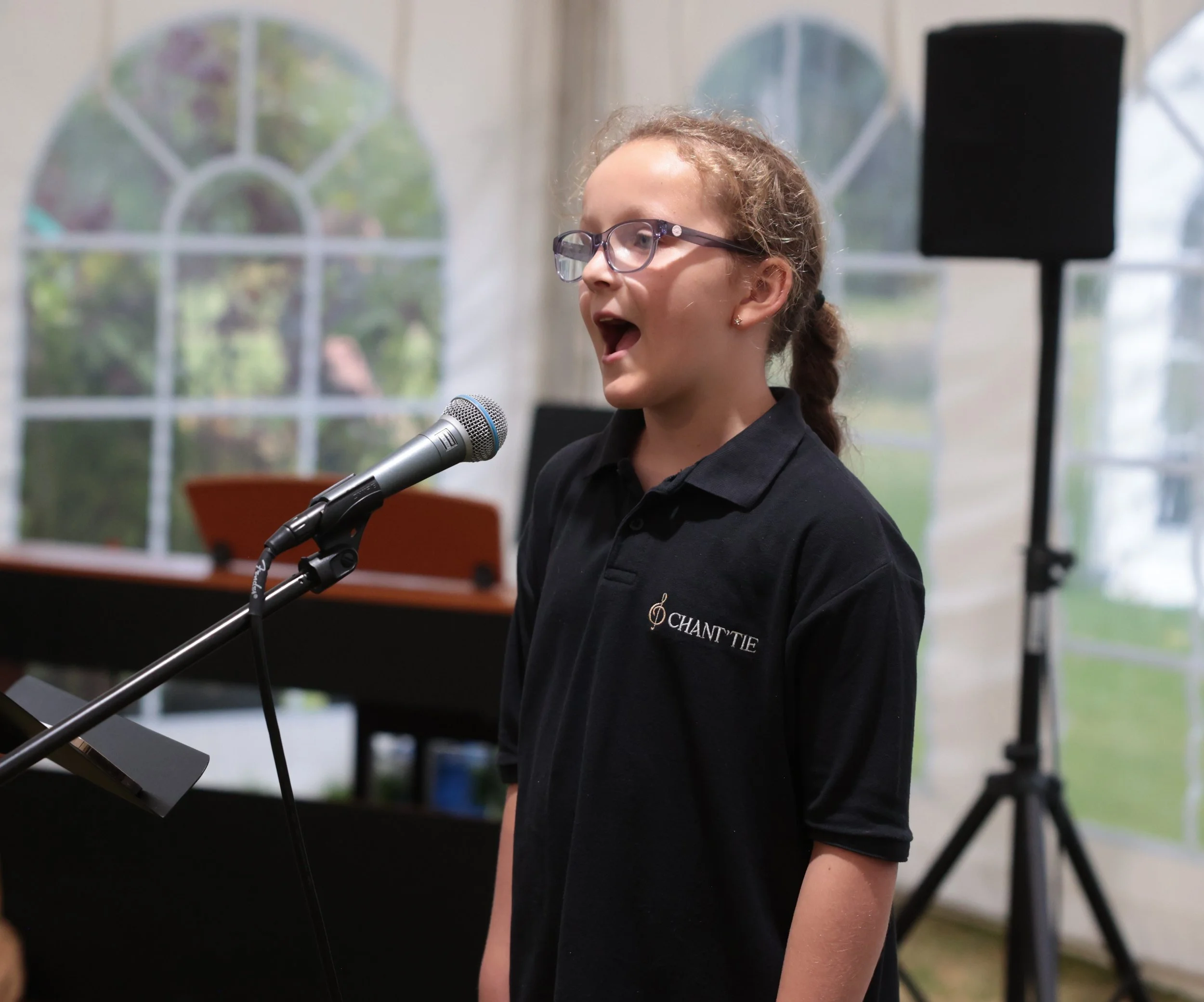 A young girl with glasses singing into a microphone inside a room with large windows.