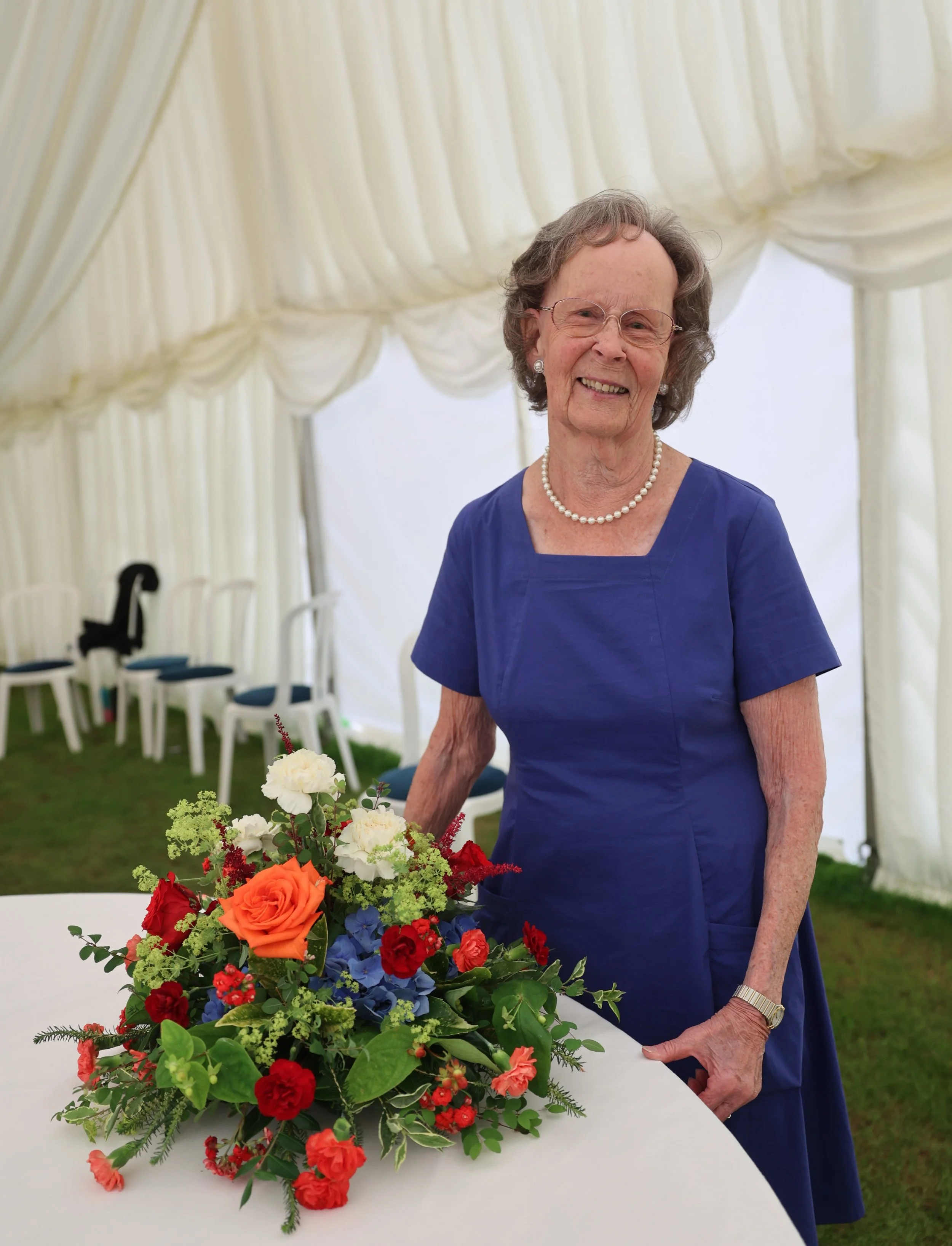 An elderly woman in a blue dress and pearl necklace smiling at a table with a large bouquet of colorful flowers, set inside a white tent with chairs in the background.
