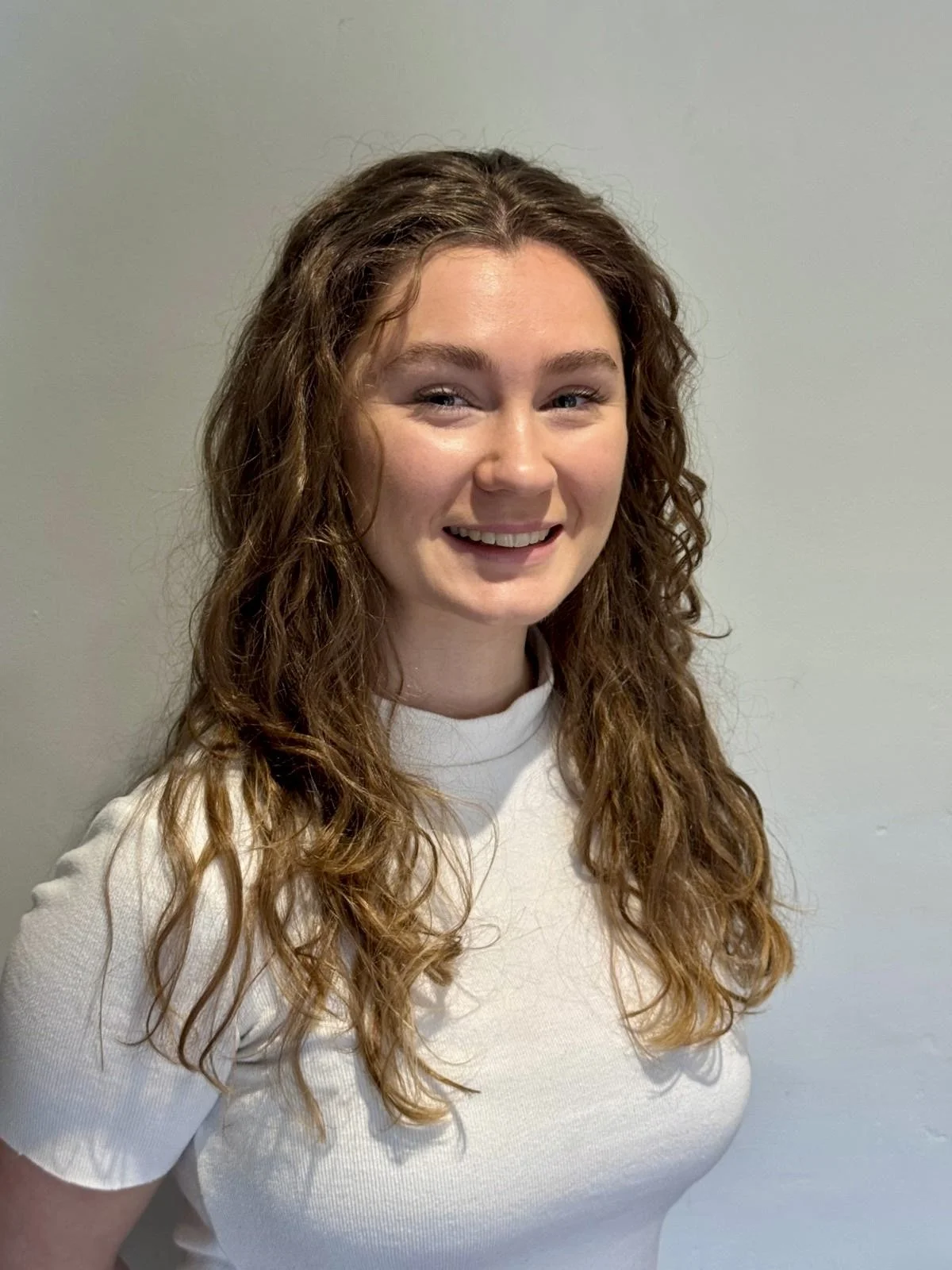 A woman with long, curly brown hair, smiling in front of a plain gray wall, wearing a white t-shirt.