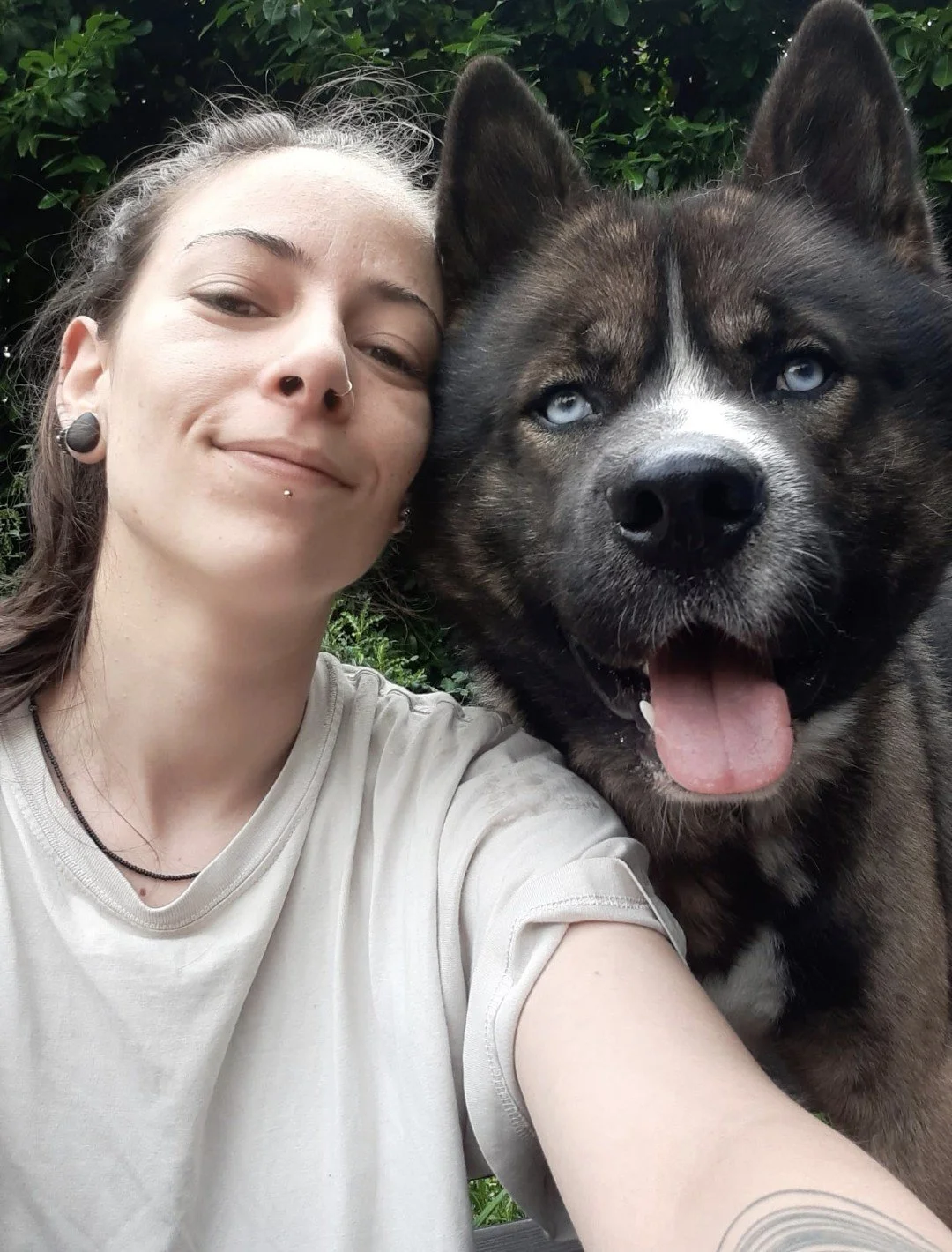 Une femme souriante posant avec un chien husky à la fourrure brune et noire, observant l'objectif avec les yeux bleus, en extérieur avec des feuillages en arrière-plan.