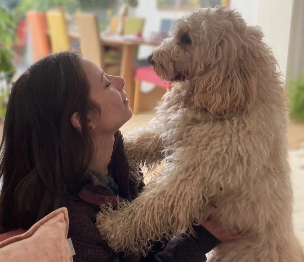 Une femme souriante face à un grand chien dun couleur beige bouclé, dans un intérieur domestique.