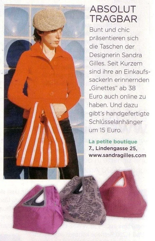 Sandra Gilles, während sie eine gestreifte Einkaufstasche aus ihrer Kollektion hält. Im Vordergrund sind drei weitere handgefertigte Handtaschen in Pink, Lila und Lila mit schwarzem Muster zu sehen.