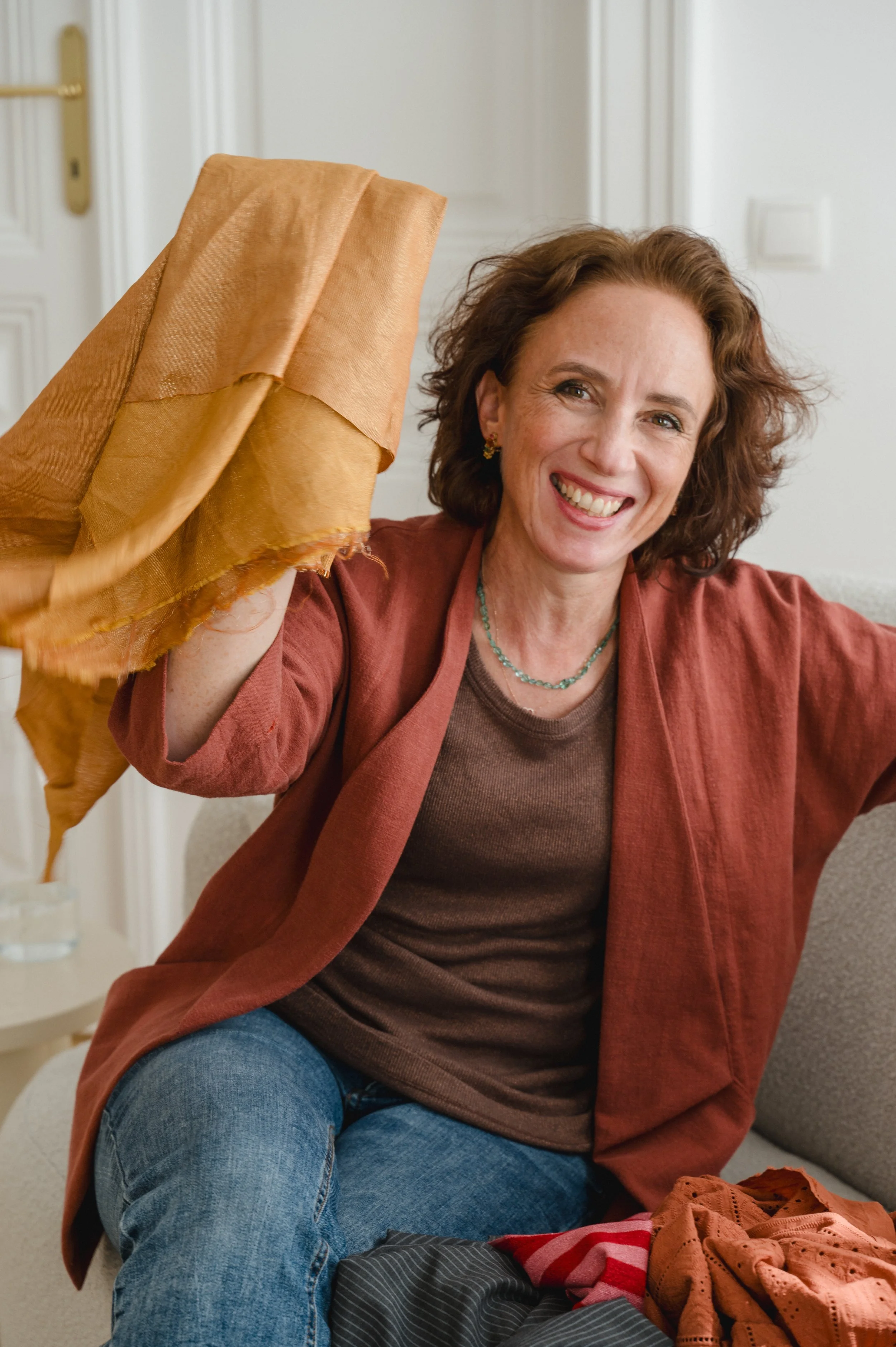 Coole, authentische Frau über 50 sitzt auf einem Sofa, lacht und hält einen Stoff in der Hand.