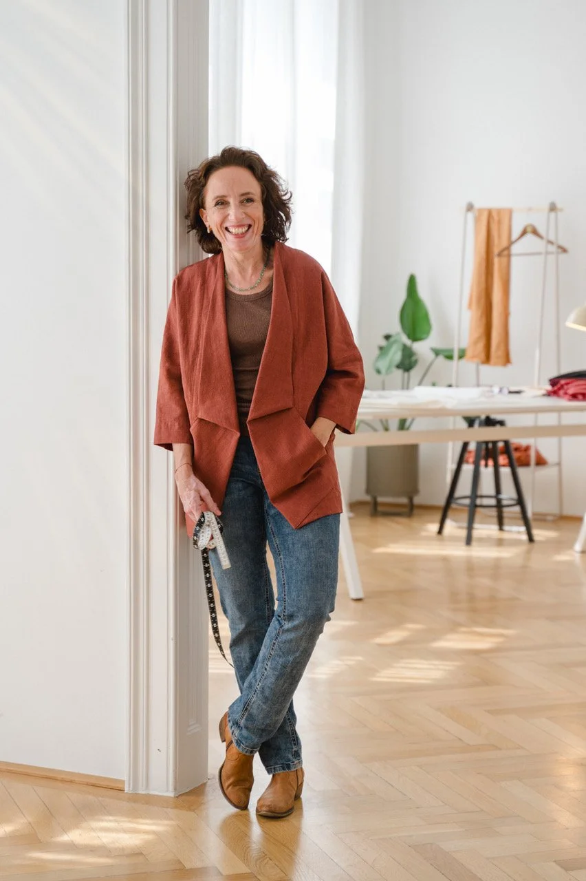 Eine lachende Frau steht in einem hellen Raum, lehnt an einer Türrahmen, trägt ein braunes Oberteil, eine rotbraune Jacke, blaue Jeans und braune Stiefeletten.