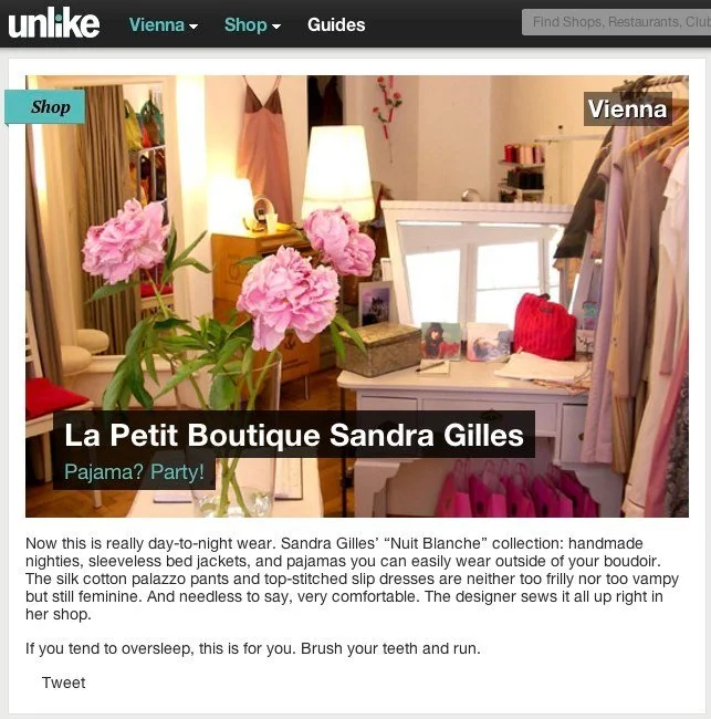 Ein Innenraum eines kleinen Modegeschäfts mit rosa Pfingstblumen, Kleidung auf Kleiderstangen und einem Spiegel, beige und weiße Einrichtungsgegenstände.