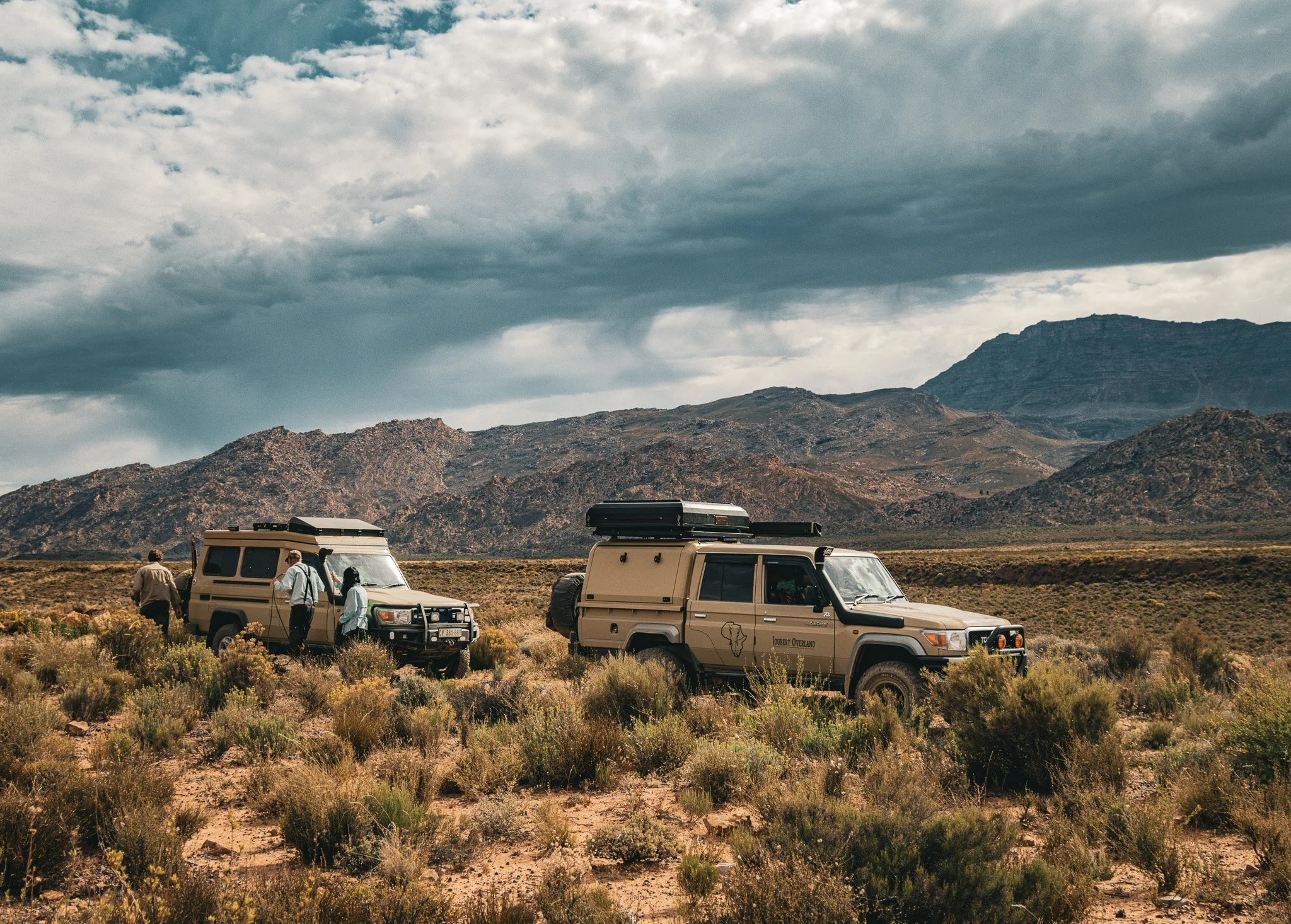 Two Land Cruiser safari vehicles on an Overland Expedition parked in a desert landscape with mountains in the background, under a cloudy sky, with people near the vehicles. The Cederberg Mountains. 