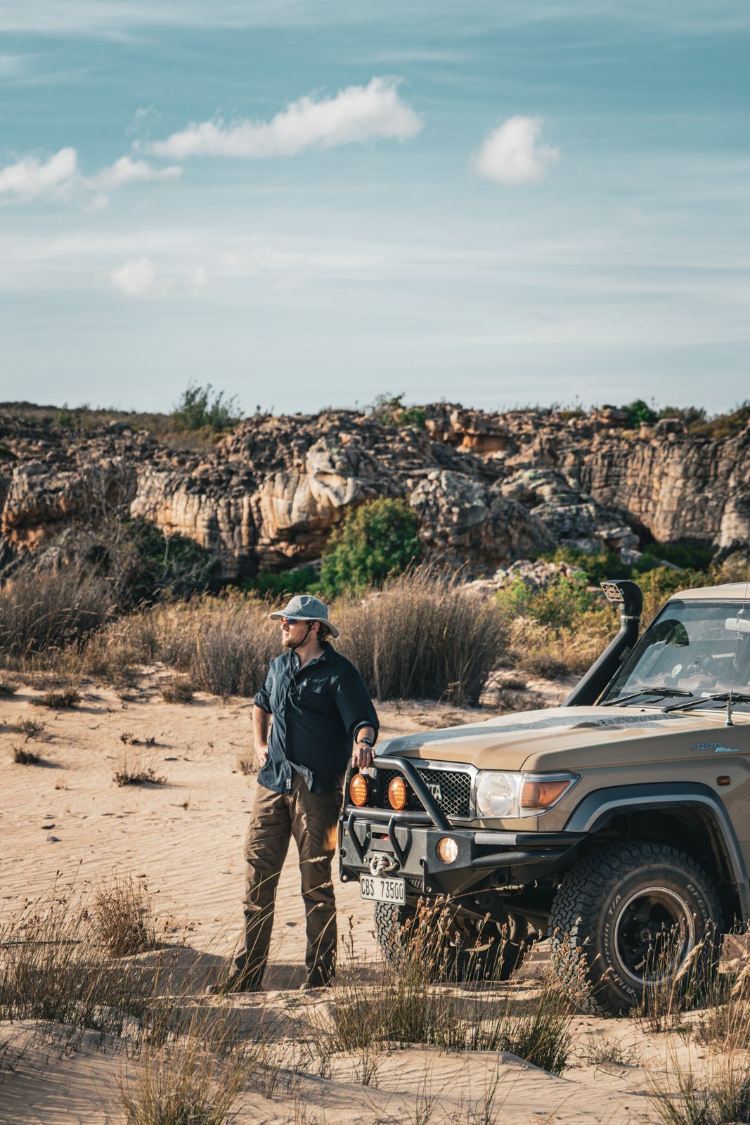 Man standing next to a beige off-road vehicle in a sandy desert landscape with rocky hills and sparse vegetation under a partly cloudy sky.