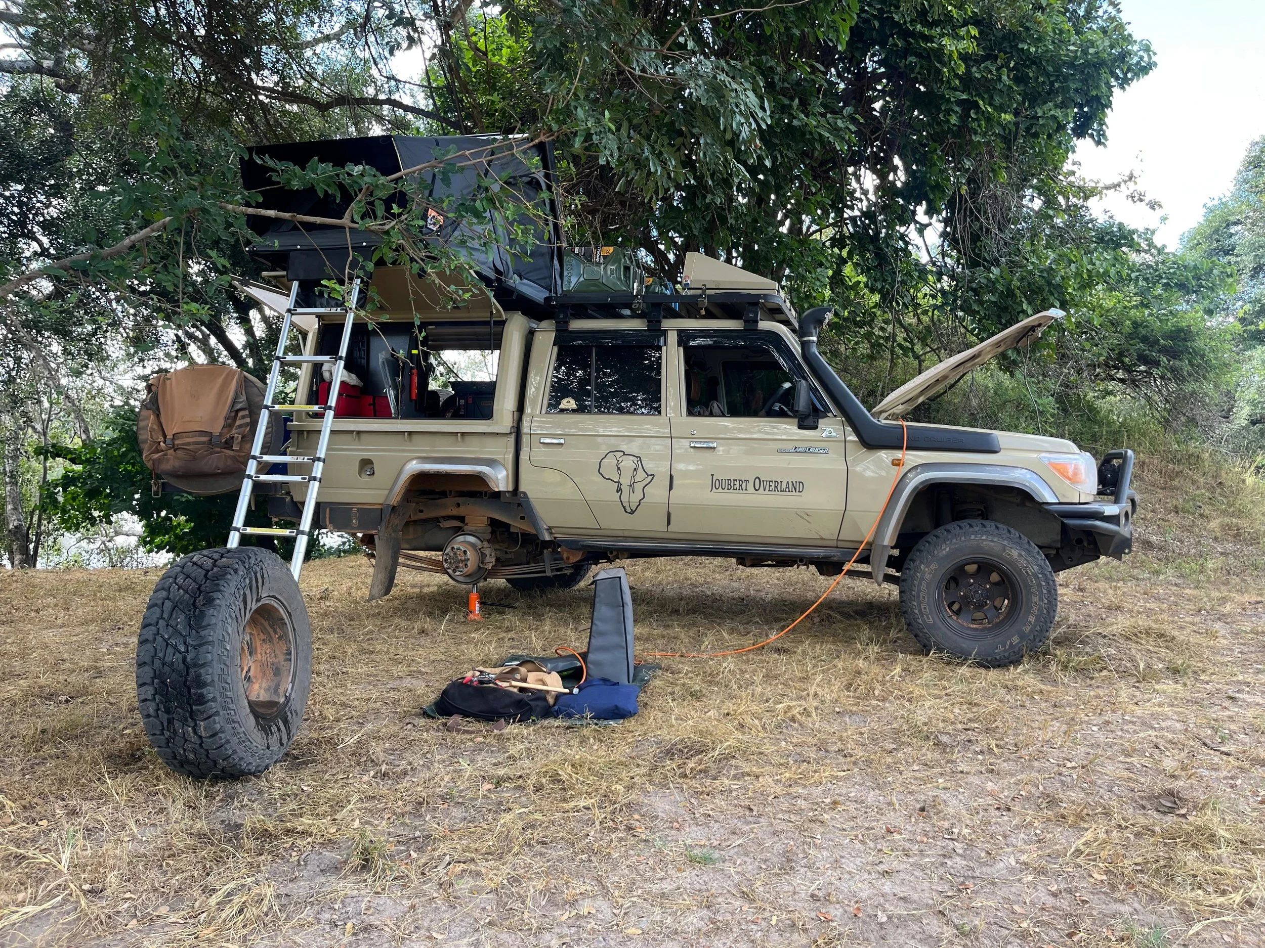 A Land Cruiser Double Cab parked on grass with open hood, surrounded by trees, camping gear on roof and ground, including a ladder, bag, and various supplies. All with the rear being repaired. 