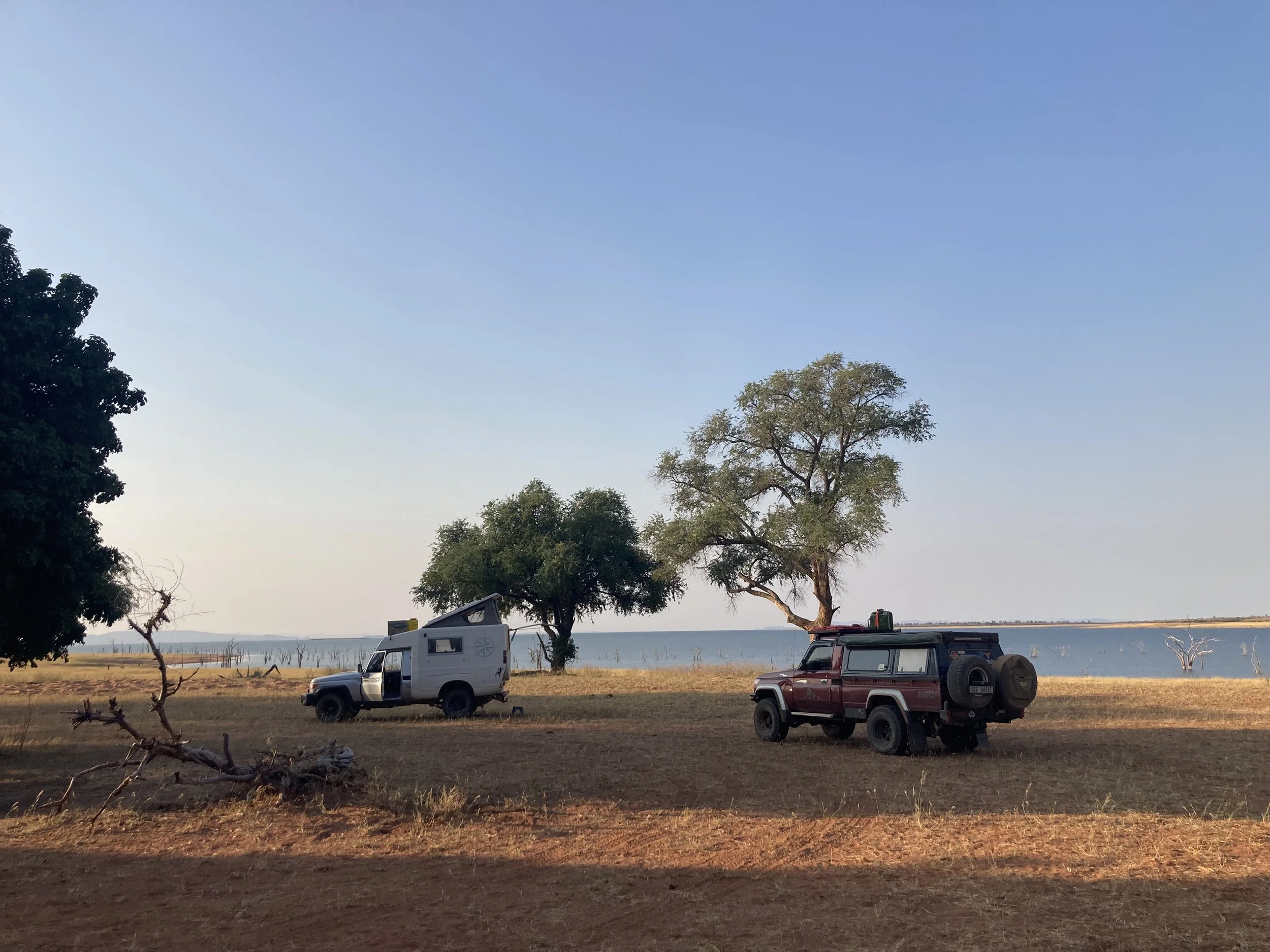 Two off-road vehicles parked on a flat, dry grassy area near a large body of water, with trees and a clear blue sky in the background.