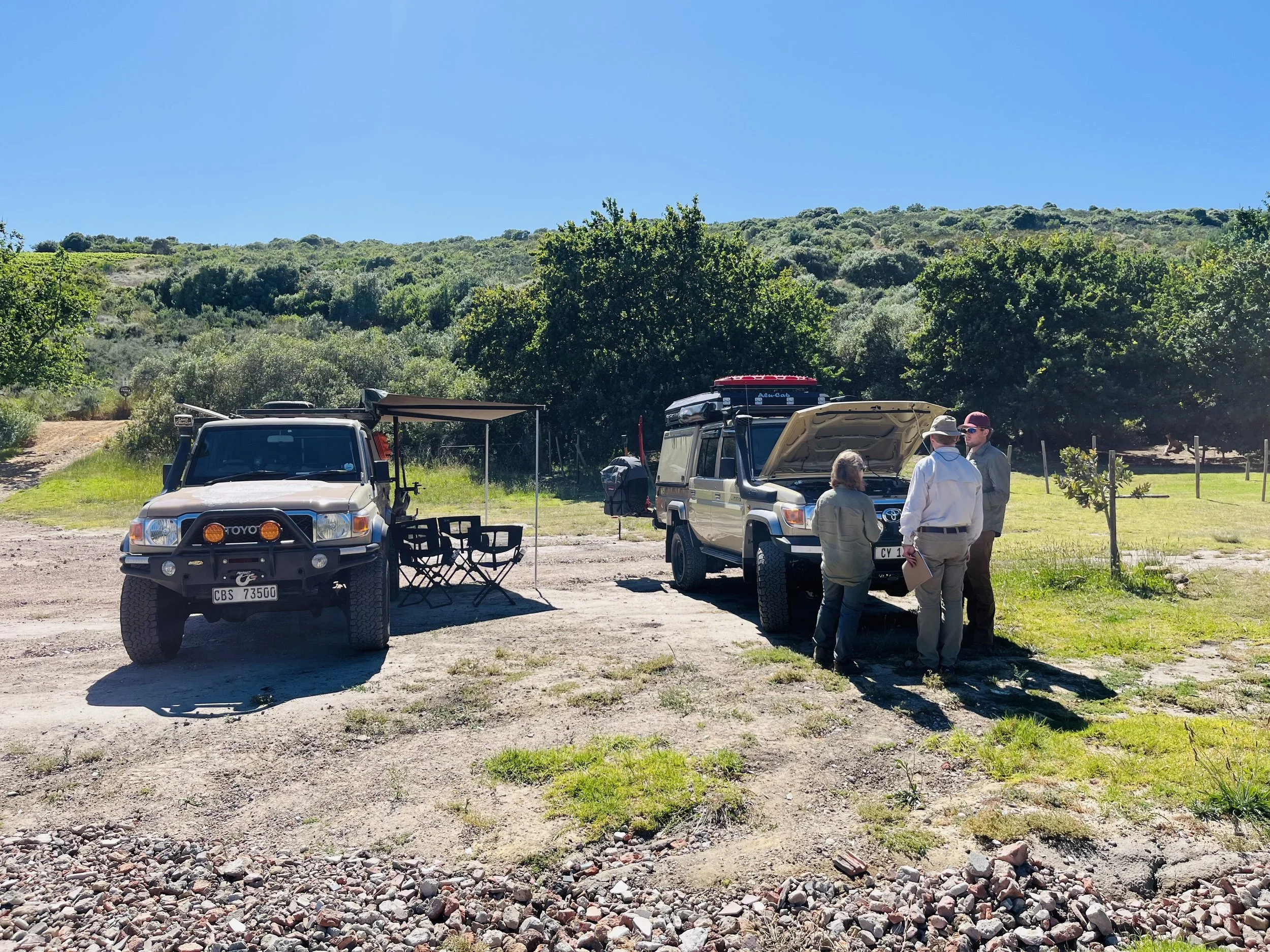 Three people standing near a Land Cruiser 79 series with its hood open in a rural outdoor setting. Another Land Cruiser with a canopy and outdoor chairs is parked nearby. The background features green trees and hills under a clear blue sky.