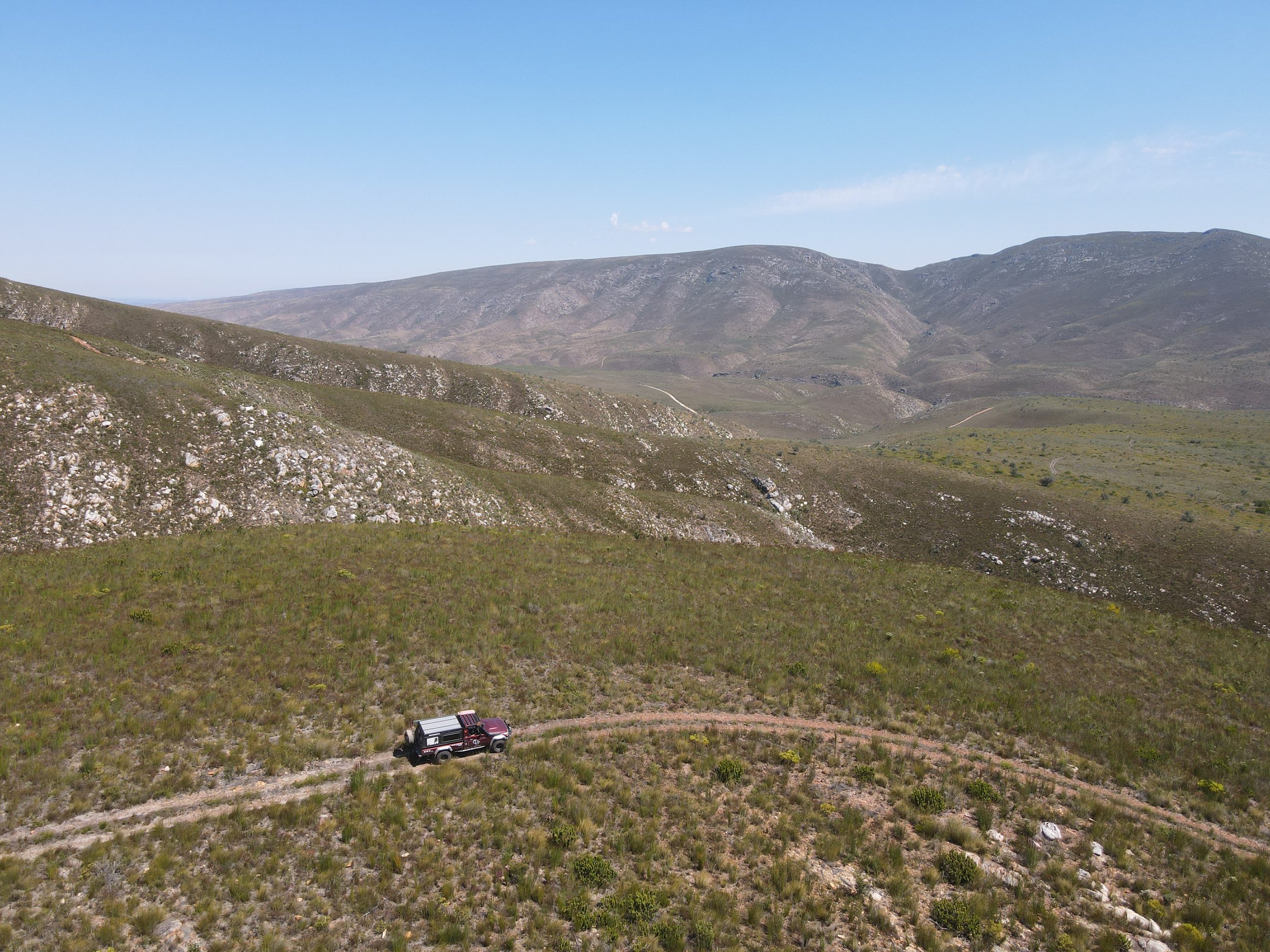 A mountainous landscape with rolling hills and sparse vegetation, featuring a lone off-road vehicle on a dirt trail.