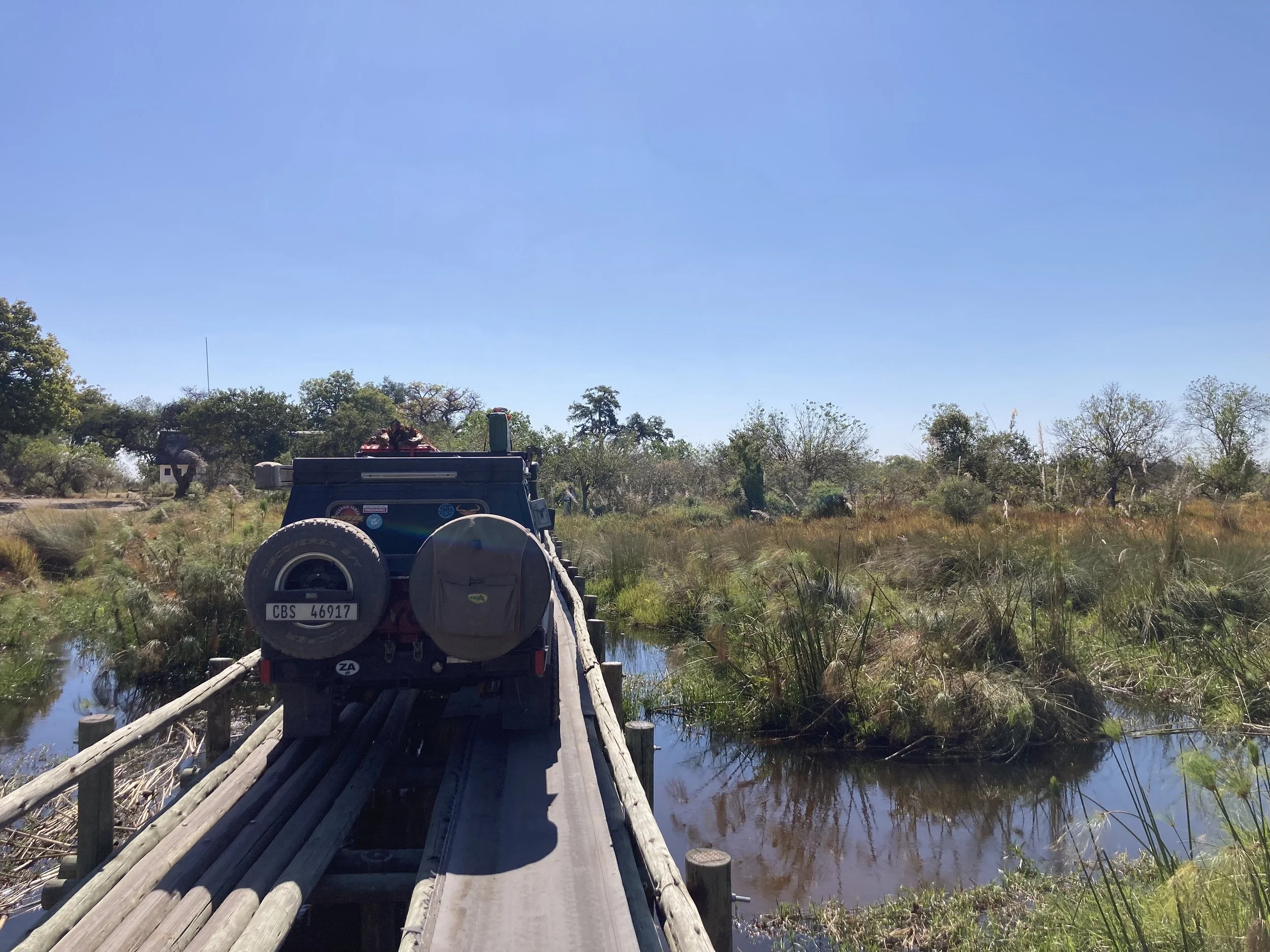 A vehicle on a narrow wooden bridge crossing over a waterway in a marshland with green grass and trees under a clear blue sky.